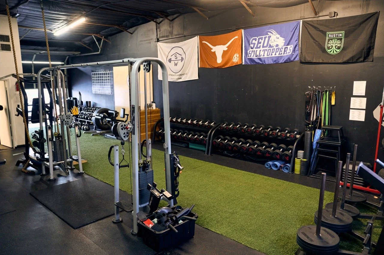 Interior of a gym with various fitness equipment, including dumbbells, weight plates, resistance bands, cable machine, and flags on the wall.