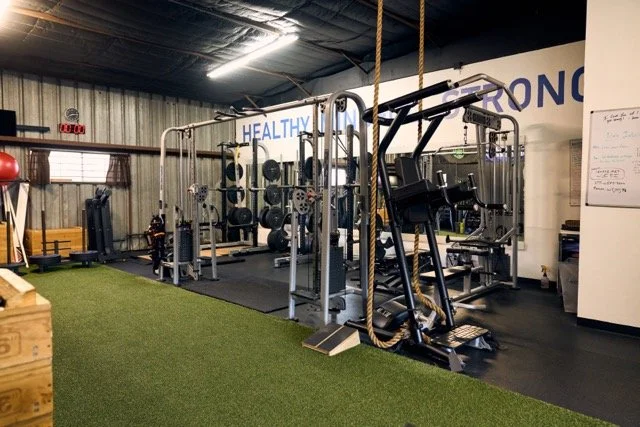 Inside a gym featuring weightlifting equipment and a climbing rope, with a sign reading 'HEALTHY. MINDFUL. STRONG.' on the wall.
