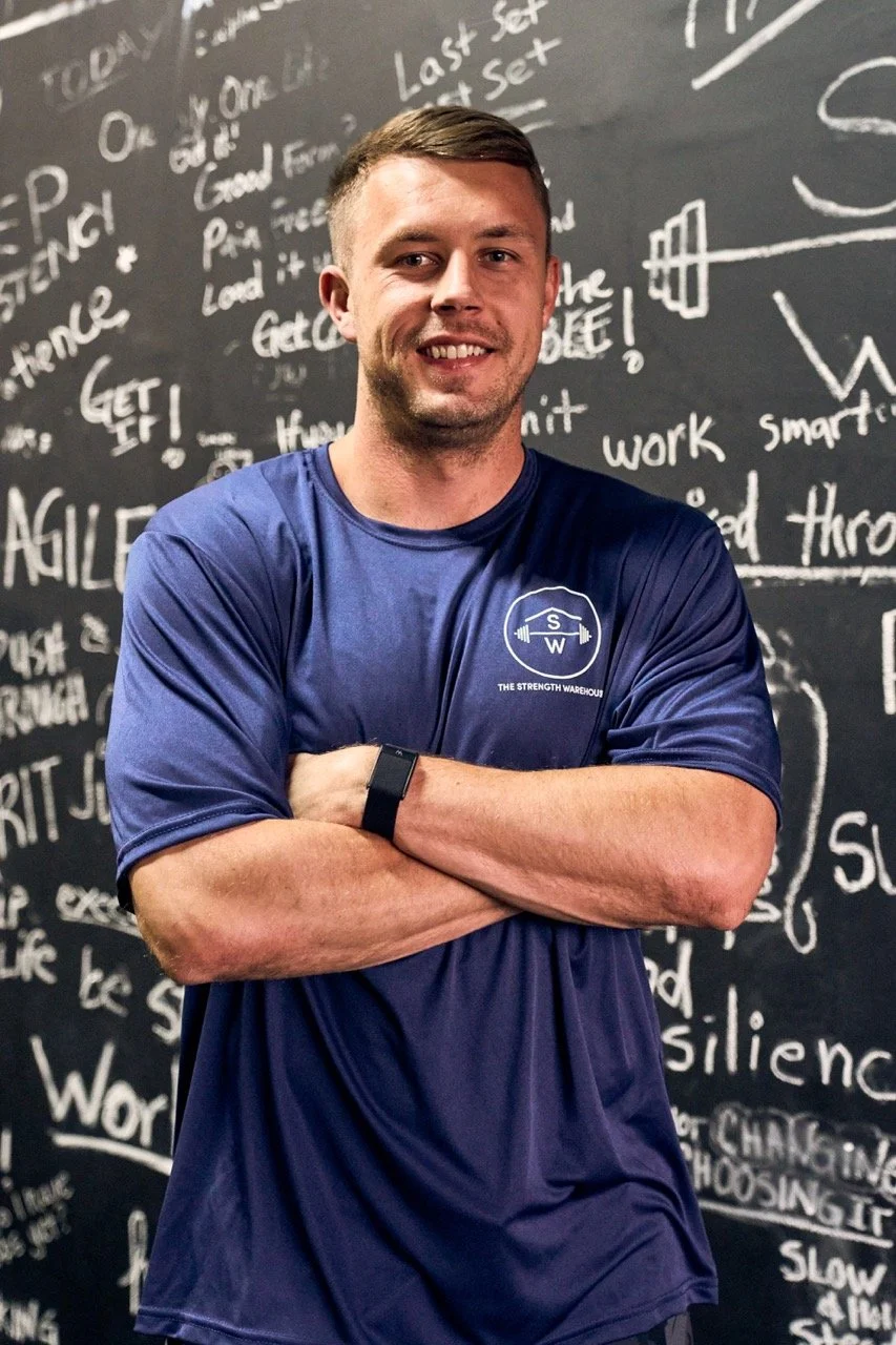 Anthony Hickey, trainer, smiling and crossing his arms, wearing a navy blue t-shirt with The Strength Warehouse logo, standing in front of the Mindfulness Wall, full of clients' intentions for their workouts