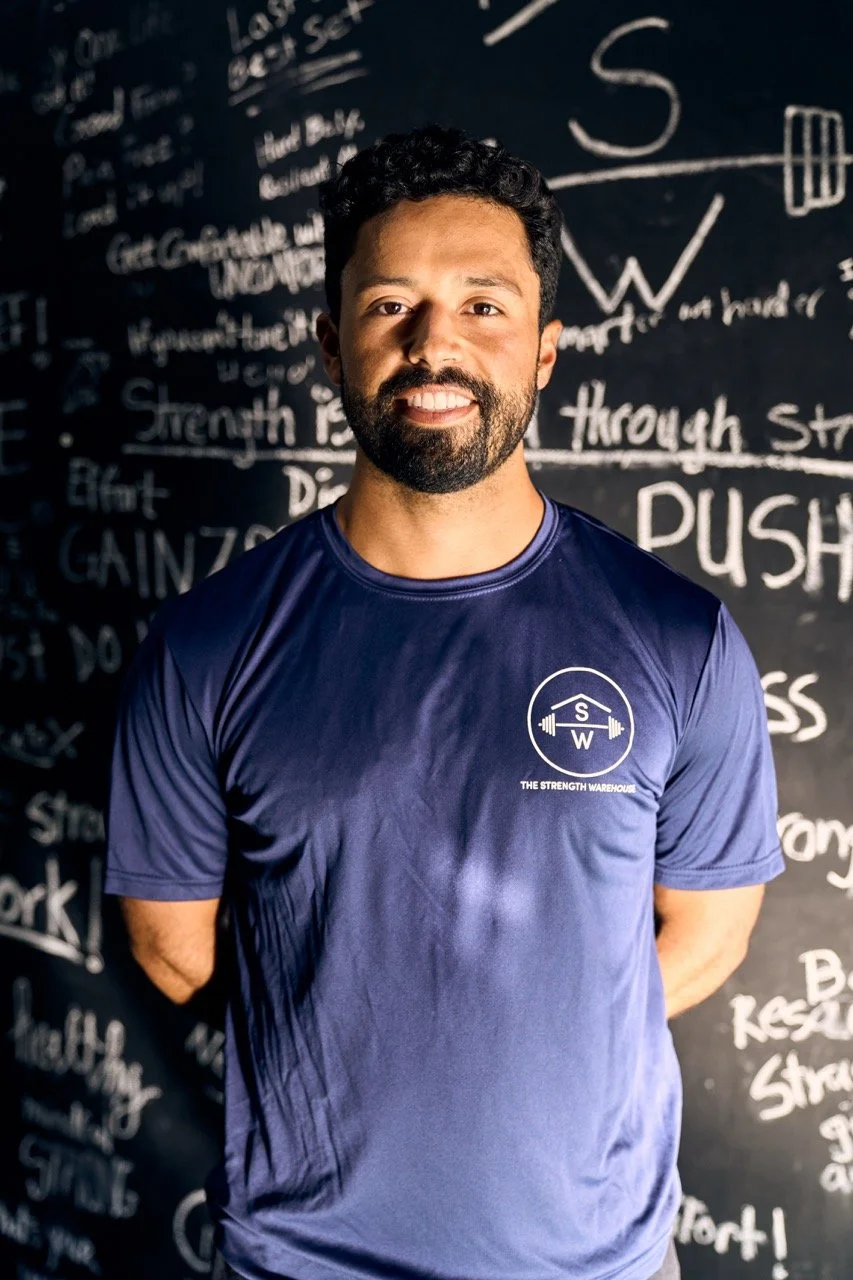 Griffin Wooden, trainer, smiling and crossing his arms, wearing a navy blue t-shirt with The Strength Warehouse logo, standing in front of the Mindfulness Wall, full of clients' intentions for their workouts