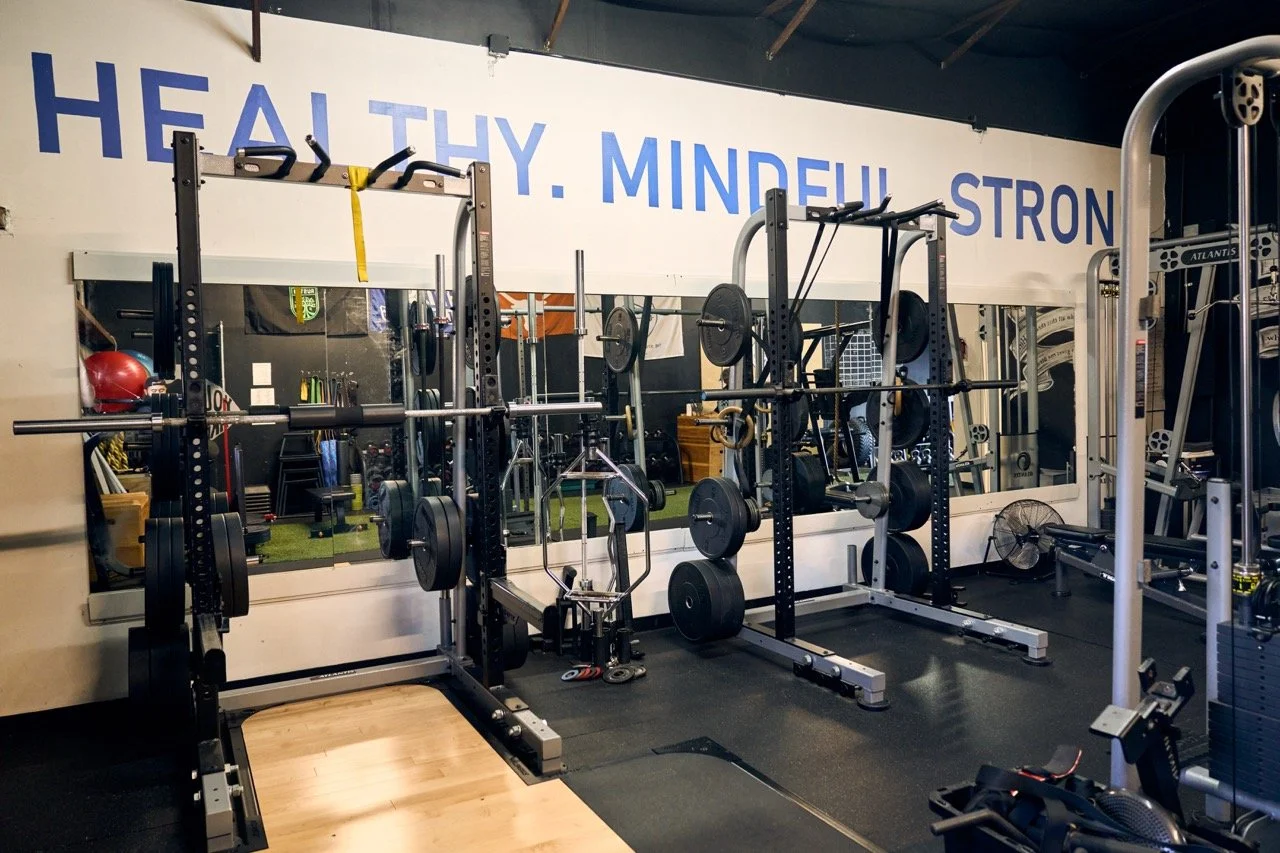 A gym with various weightlifting equipment including squat racks, barbells, weights, a small fan, and a large mirror with the words 'HEALTHY. MINDFUL. STRONG.' written above it.