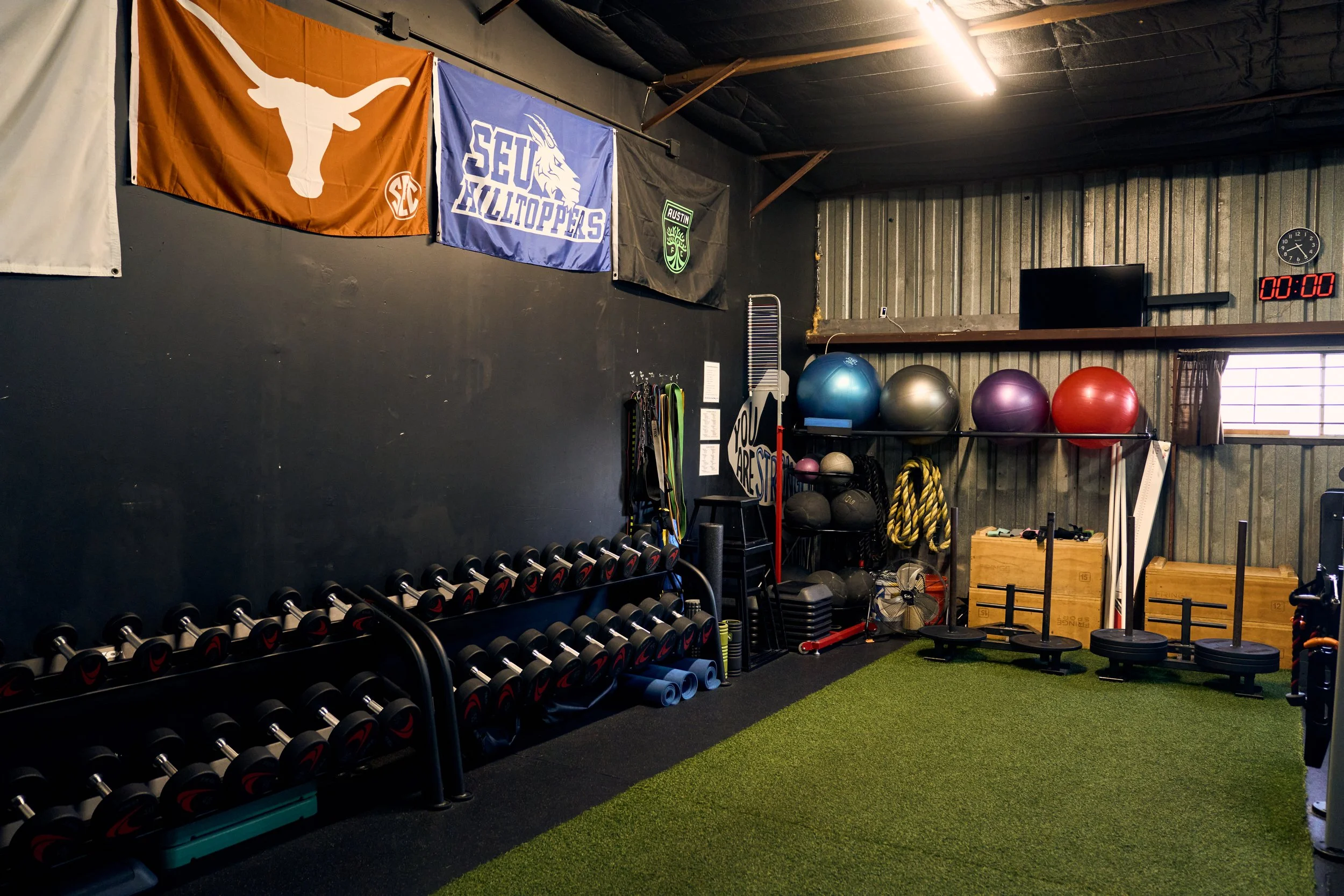 A gym with black walls and flags hanging on the wall. Equipment includes dumbbells on racks, stability balls, medicine balls, battle ropes, plyometric boxes, and a green turf mat.