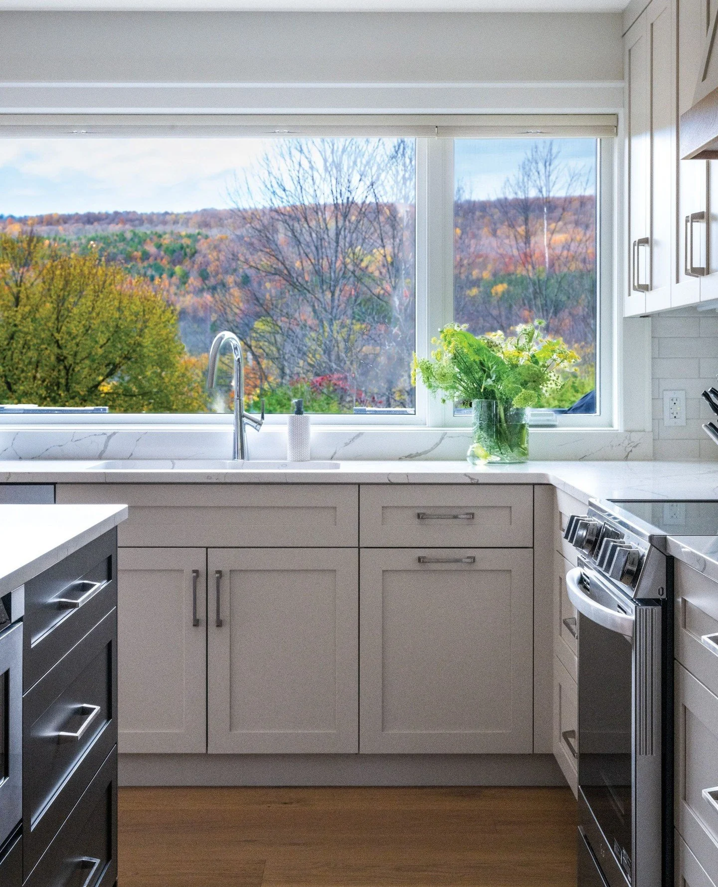 The inspiring view is captured through large windows in this redesigned kitchen in a Beaver Valley bungalow. Adding a large quartz island is ideal for casual meals with plenty of prep space and storage. Open oak shelving and the pantry are two of the