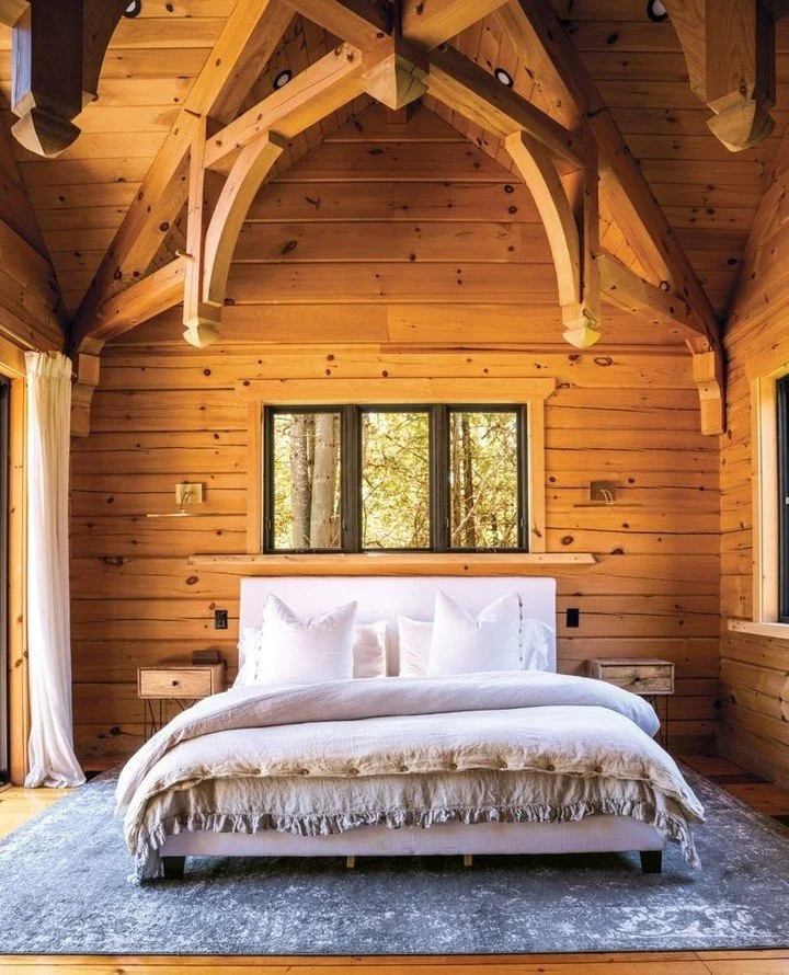The picturesque primary bedroom in this log home is tucked into the east wing for optimal privacy. It features a double hammer beam truss for a grand yet cosy look. The primary en suite was designed to feel like an at-home spa with a luxurious soaker