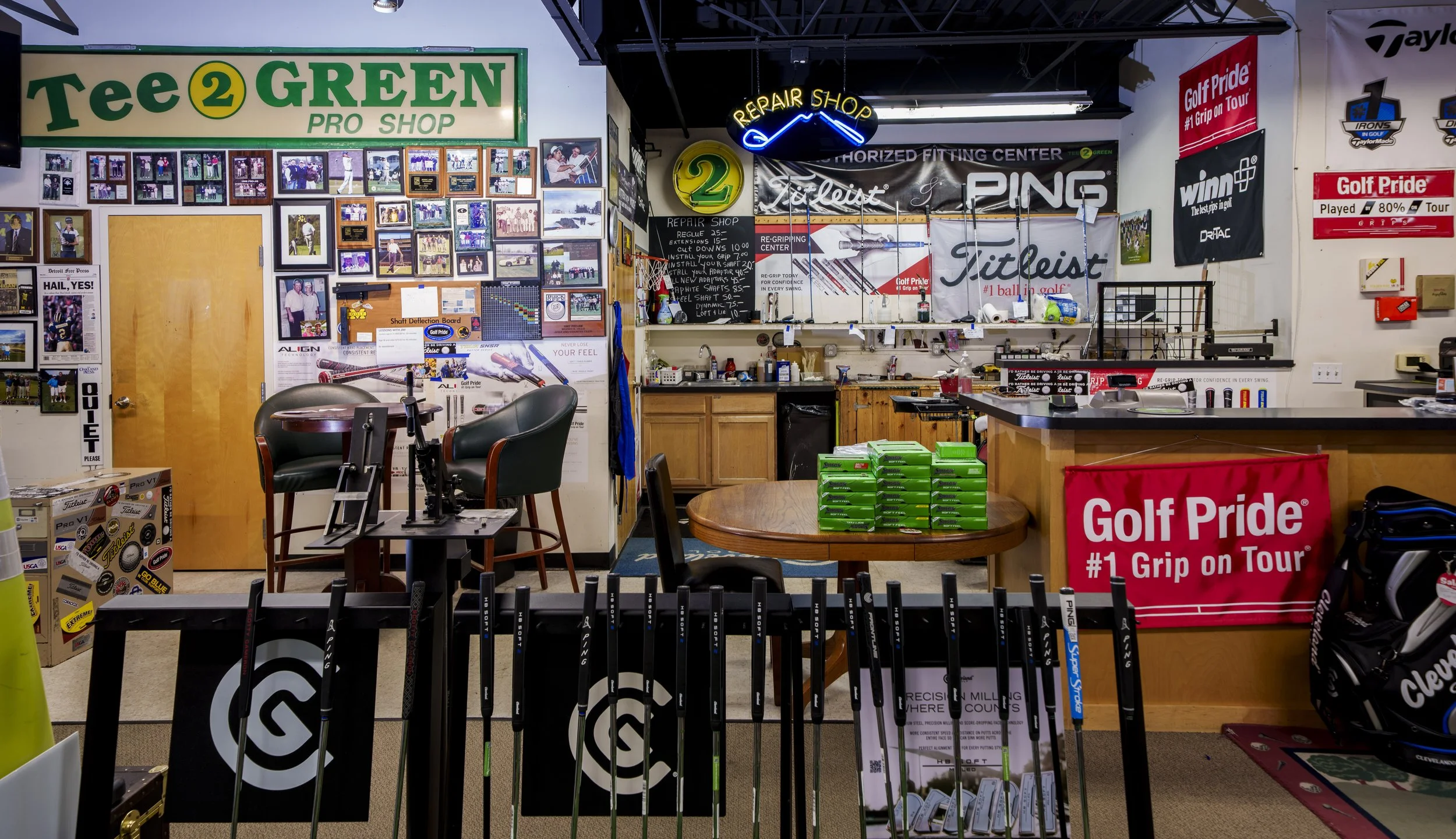 Inside a golf shop with walls covered in photos, posters, and banners related to golf. There are golf clubs on display, a table with boxes of golf balls, and signs advertising golf brands and services.