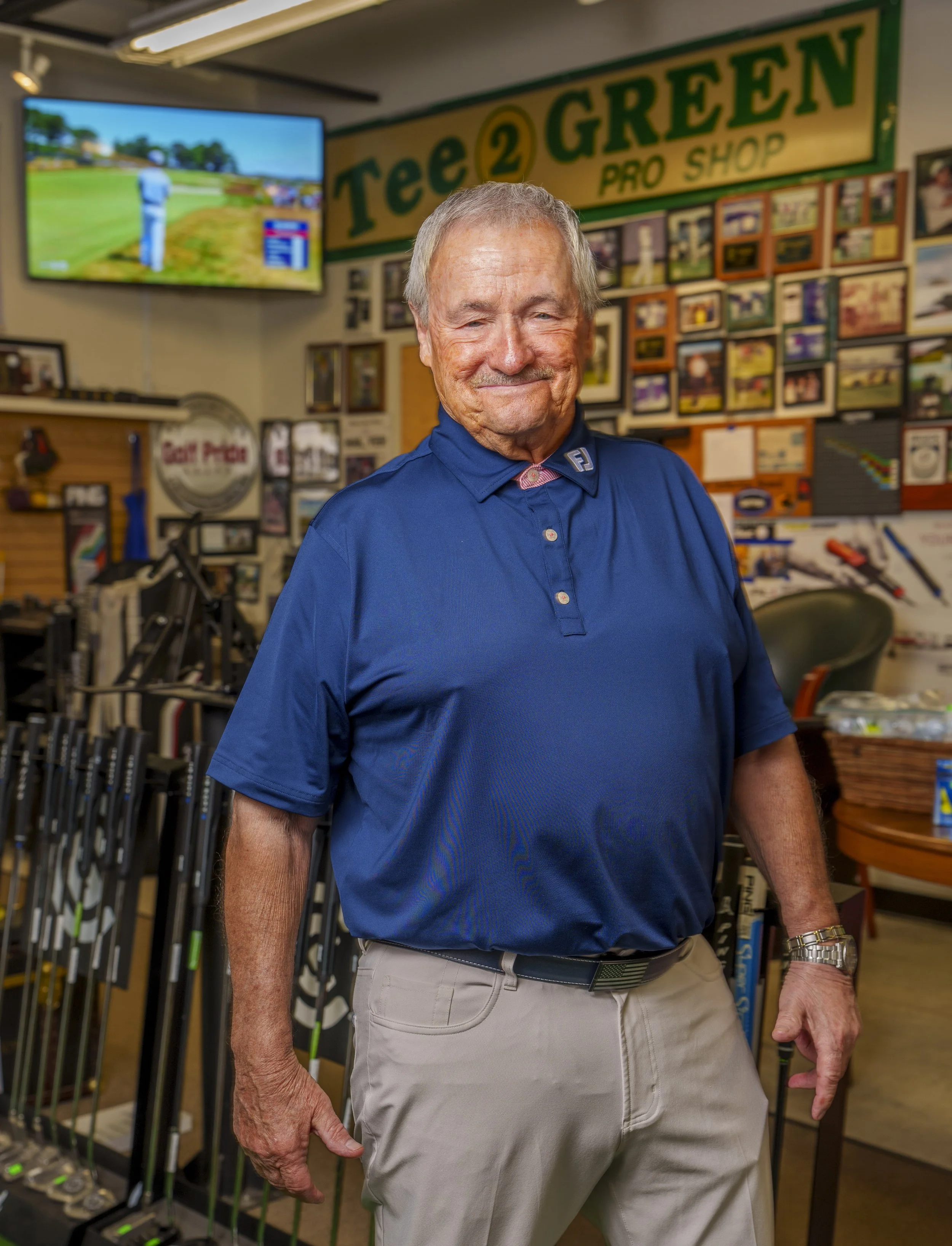 An elderly man smiling in a golf shop surrounded by golf clubs and golf-related items.