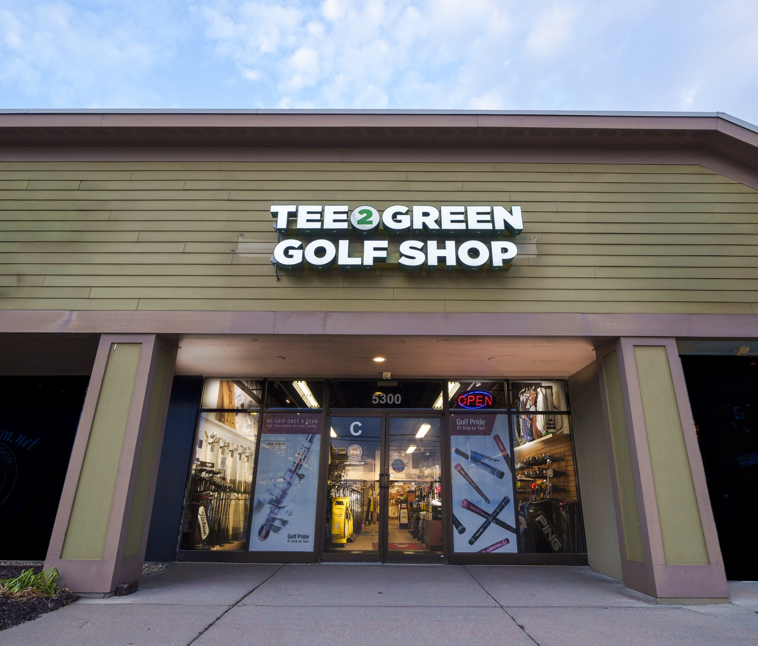 Front view of a golf shop named 'Tee 2 Green Golf Shop' with an 'OPEN' sign, showing golf clubs and accessories inside.