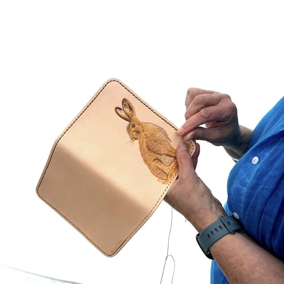 Wild Hare Stitch.jpg A6 brown hare, leather journal cover being stitched by maker. Hare is golden brown on natural background.