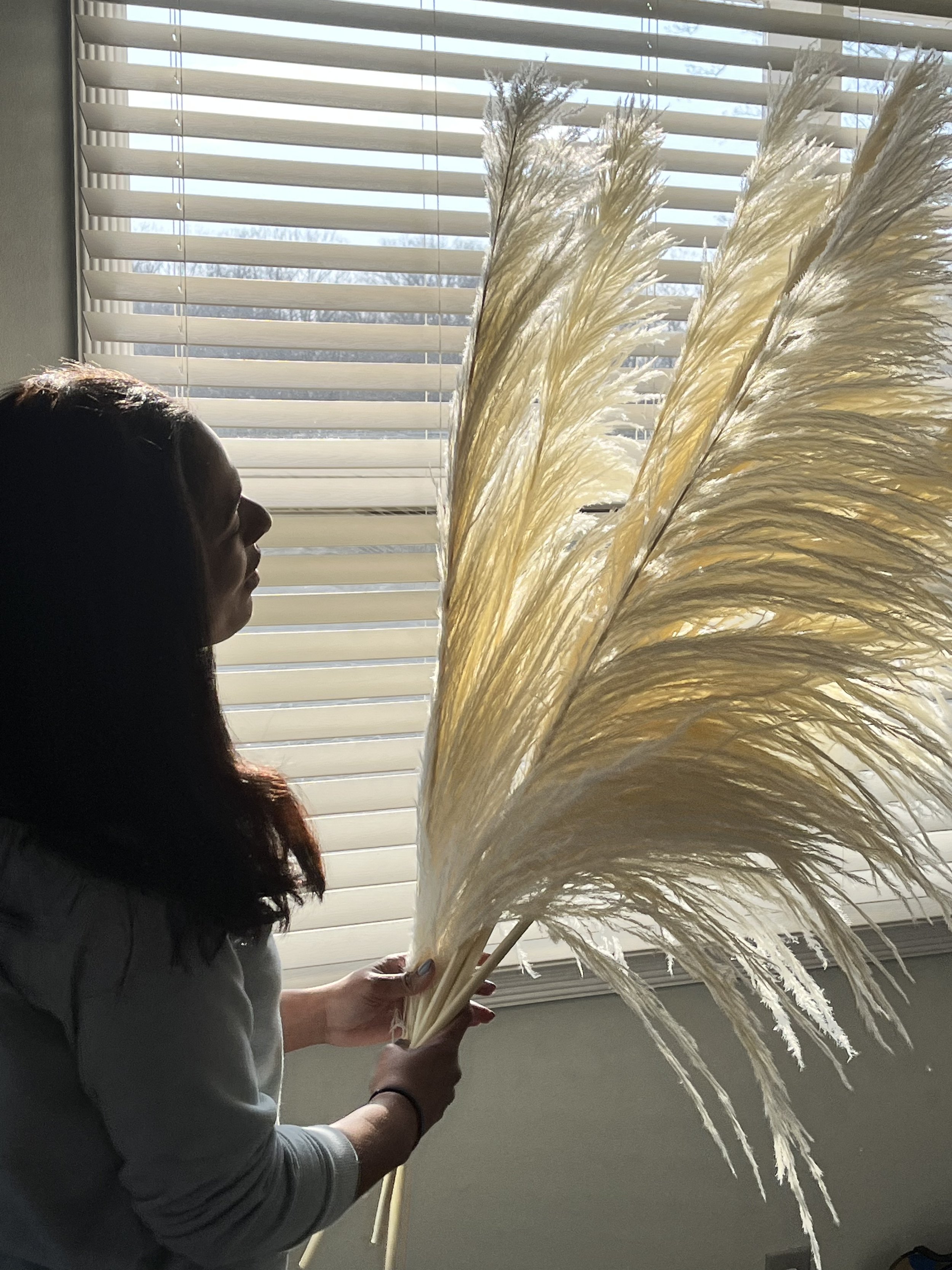 Elise with dark hair holding a large bunch of dried pampas grass by a window with horizontal blinds, sunlight filtering through.