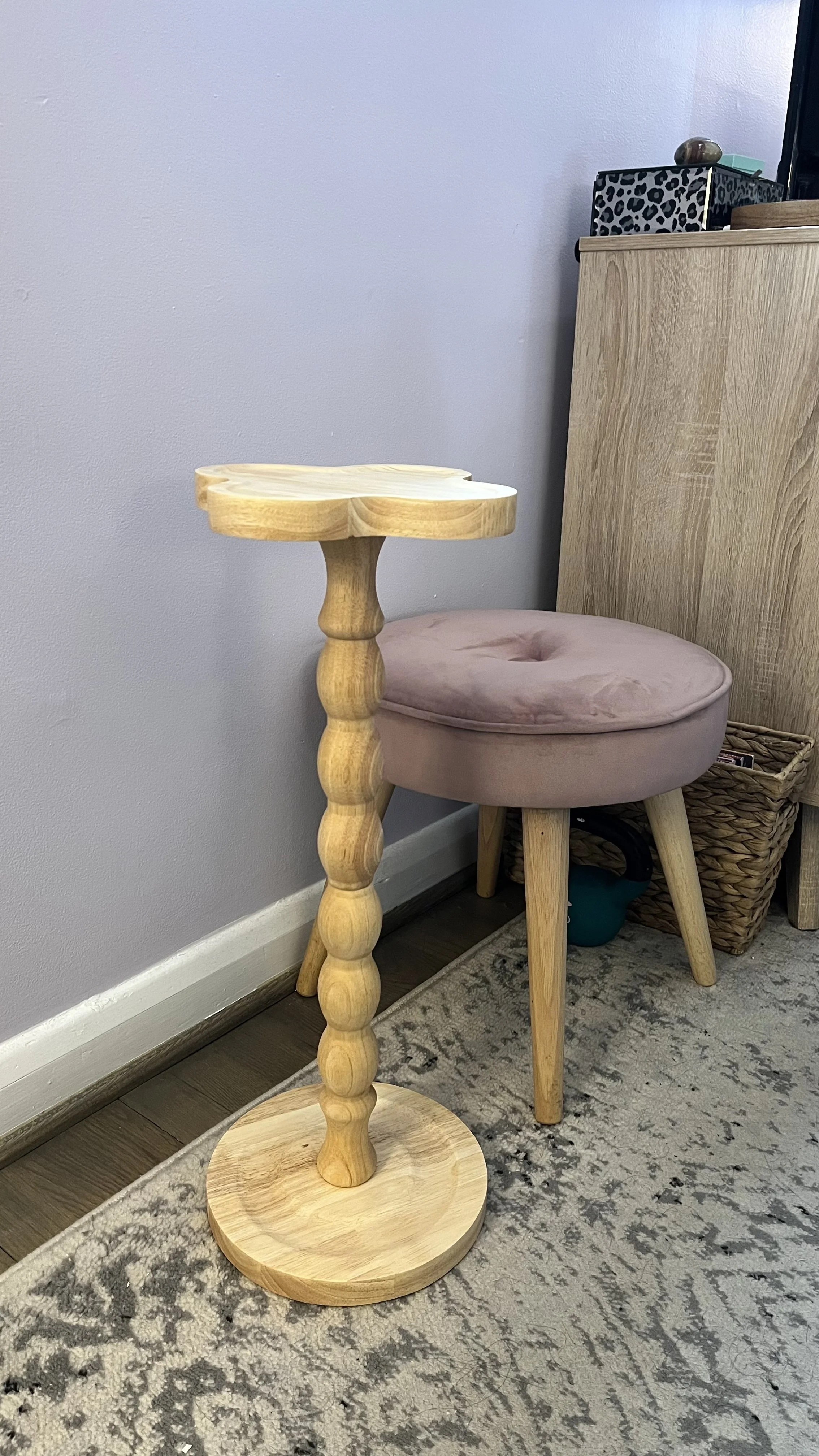 A wooden side table with a round base and a carved, spindle-style stand, positioned next to a pink cushioned stool with three legs, against a light gray wall and a wooden cabinet.