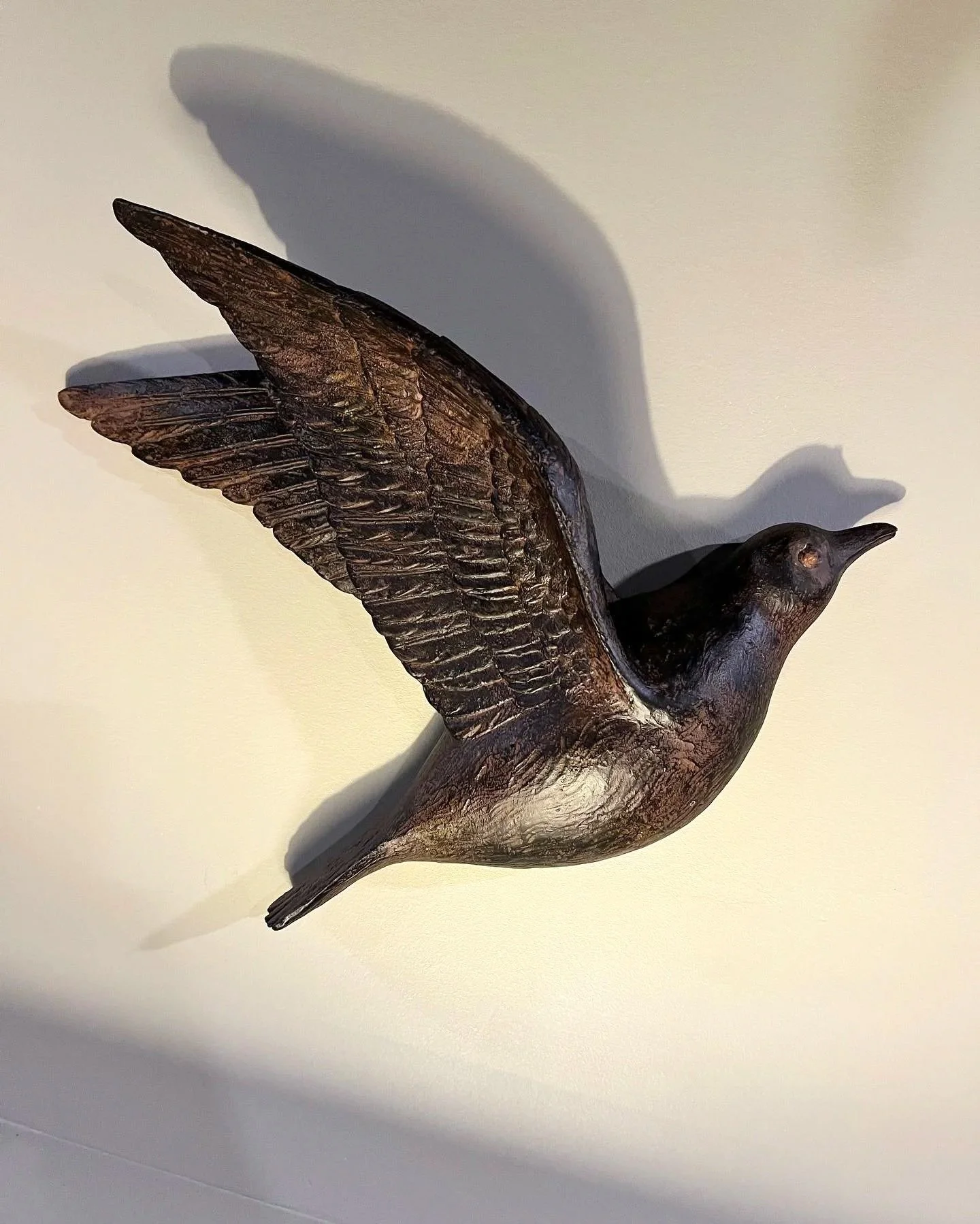 Metal sculpture of a bird in flight with outstretched wings on a light-colored background.