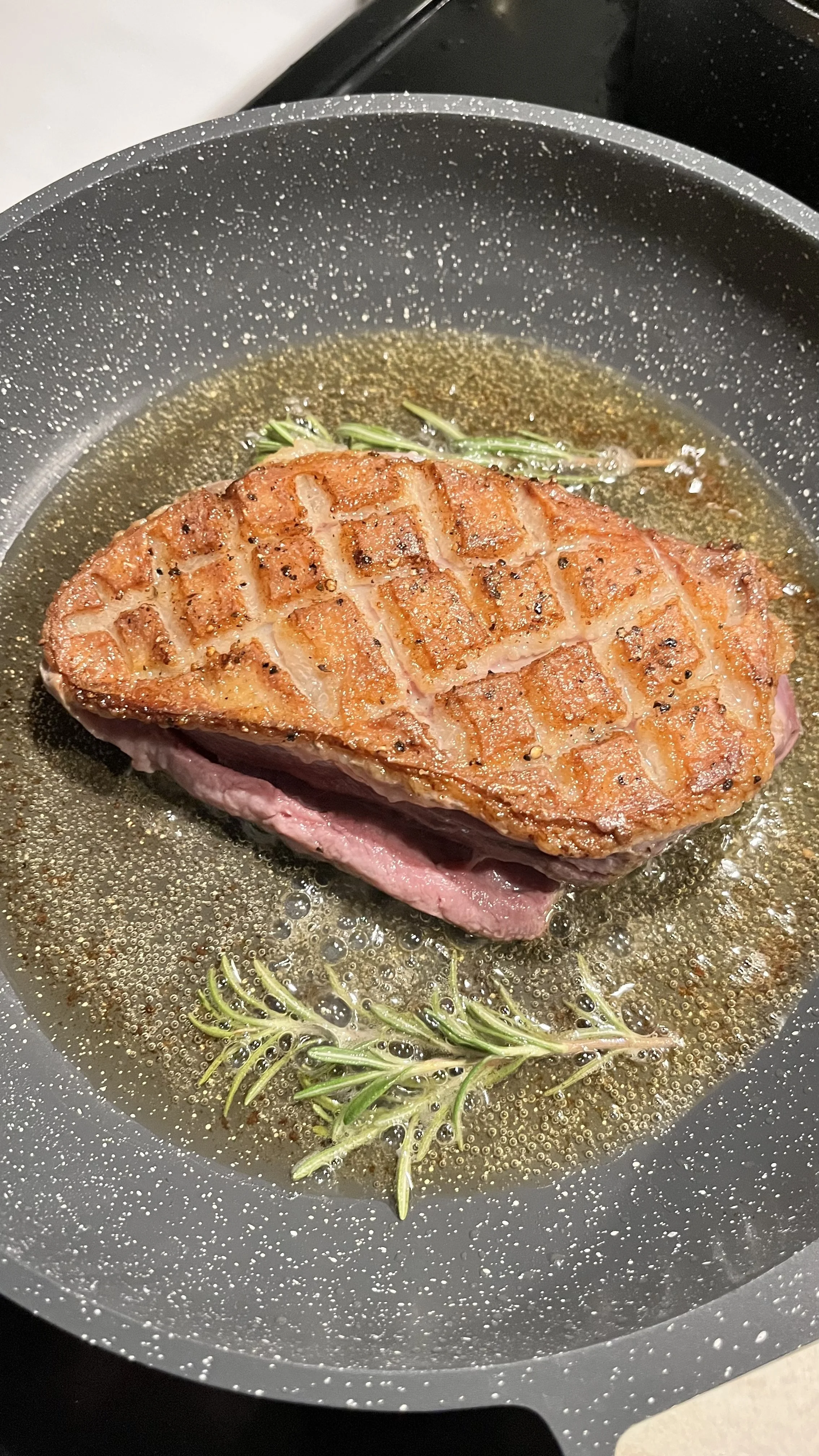 A piece of steak being seared in a pan with herbs, likely rosemary, and rendered fat.