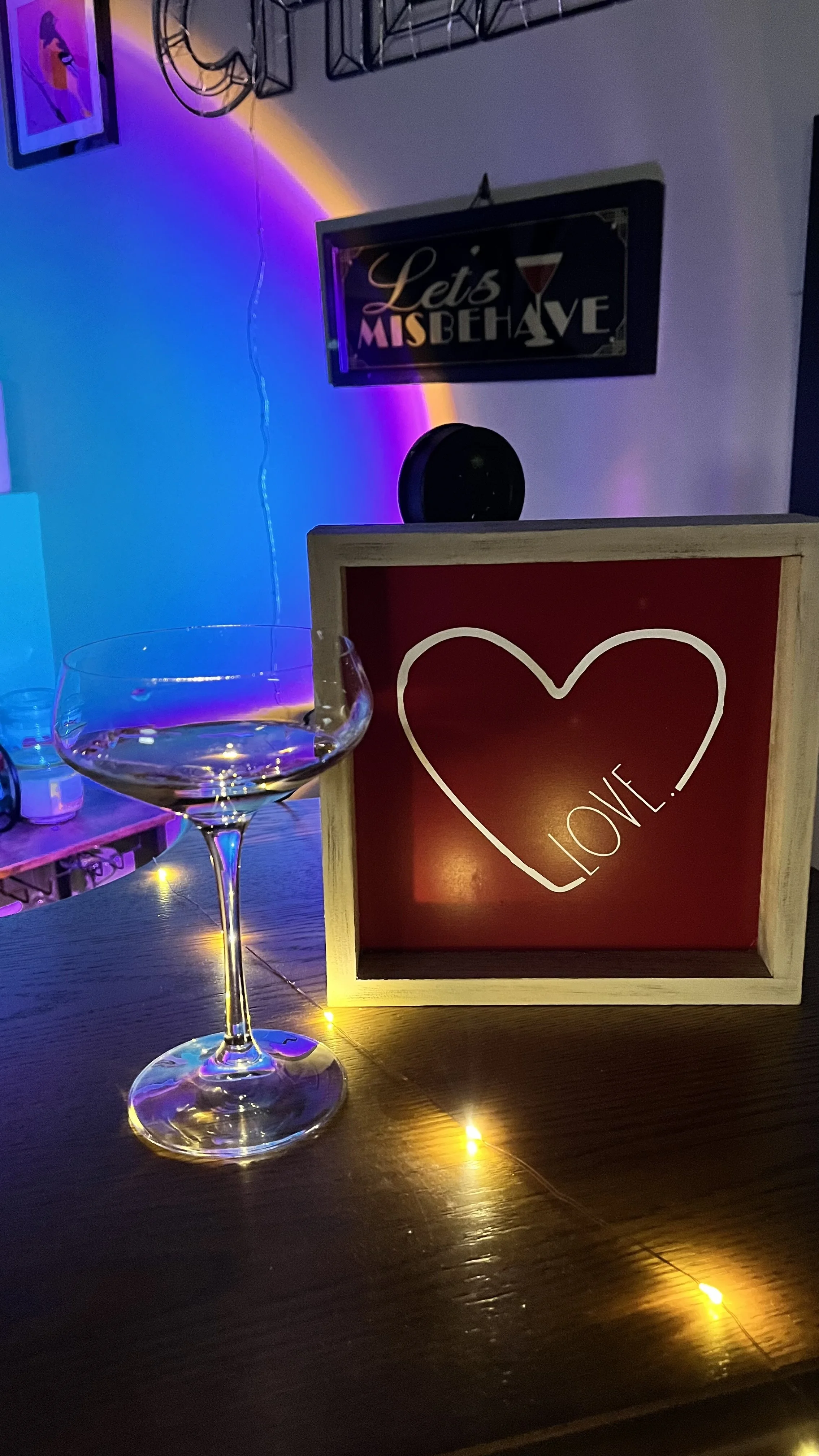 A bar setting with a glass of wine, a red and white sign that says 'LOVE' with a heart outline, and a neon-lit sign that says 'Let's Misbehave.' There are string lights and a purple LED light creating a colorful ambiance.