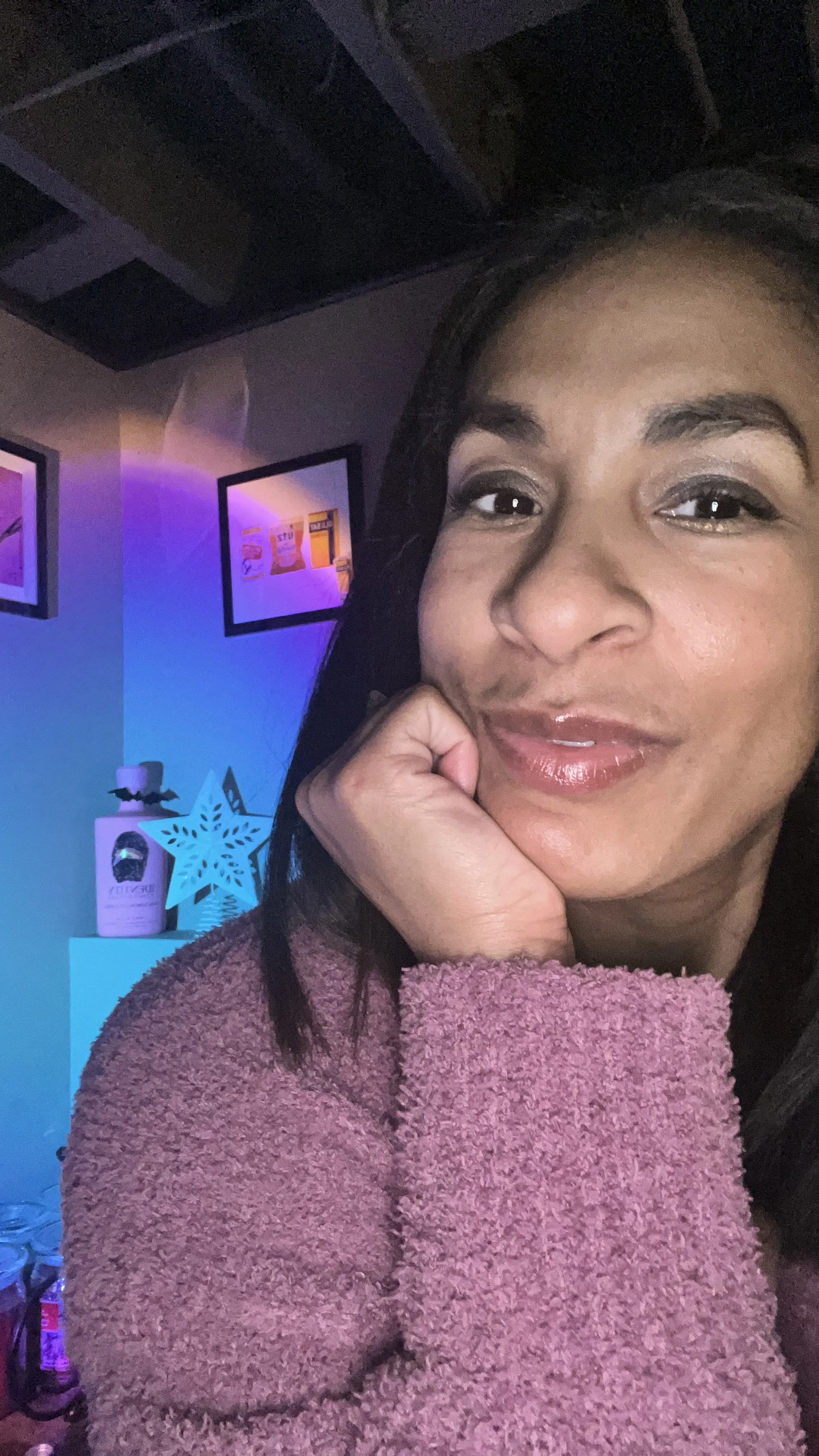 Elise with dark hair and a pink fuzzy sweater, resting her chin on her hand and smiling at the camera. In the background, there is a blue star decoration and illuminated framed pictures on the wall.