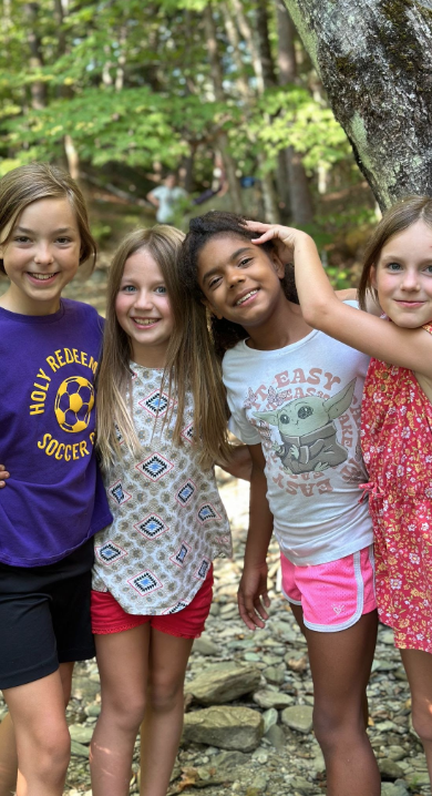  Four young people stand together smiling. They are wearing a purple soccer shirt, a patterned shirt, a "Baby Yoda" shirt, and a red floral dress. 