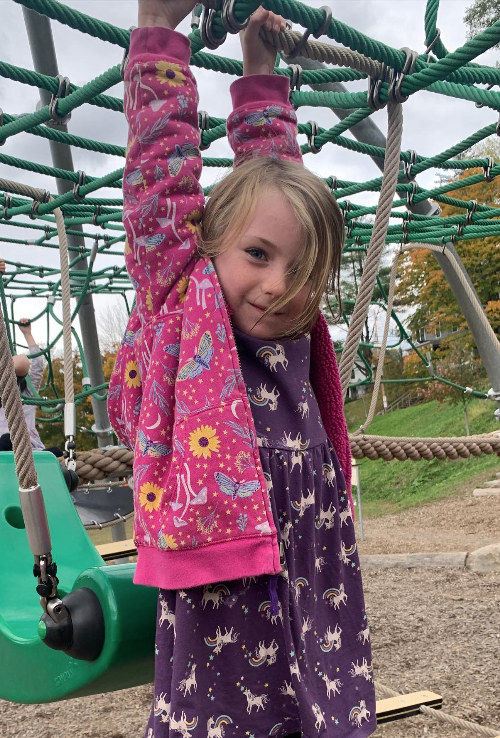  A smiling child with blonde hair hangs from a rope climbing structure on a playground. They are wearing a purple dress with unicorns and a pink floral hoodie. 