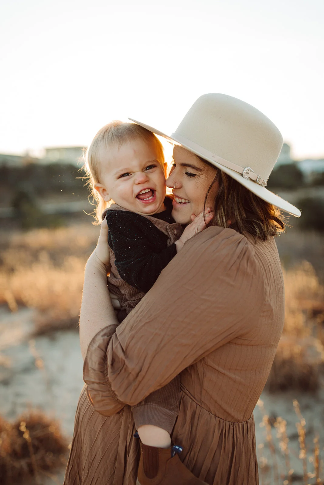 A woman wearing a beige wide-brim hat and tan dress holding a laughing toddler with a nose piercing outdoors during sunset.