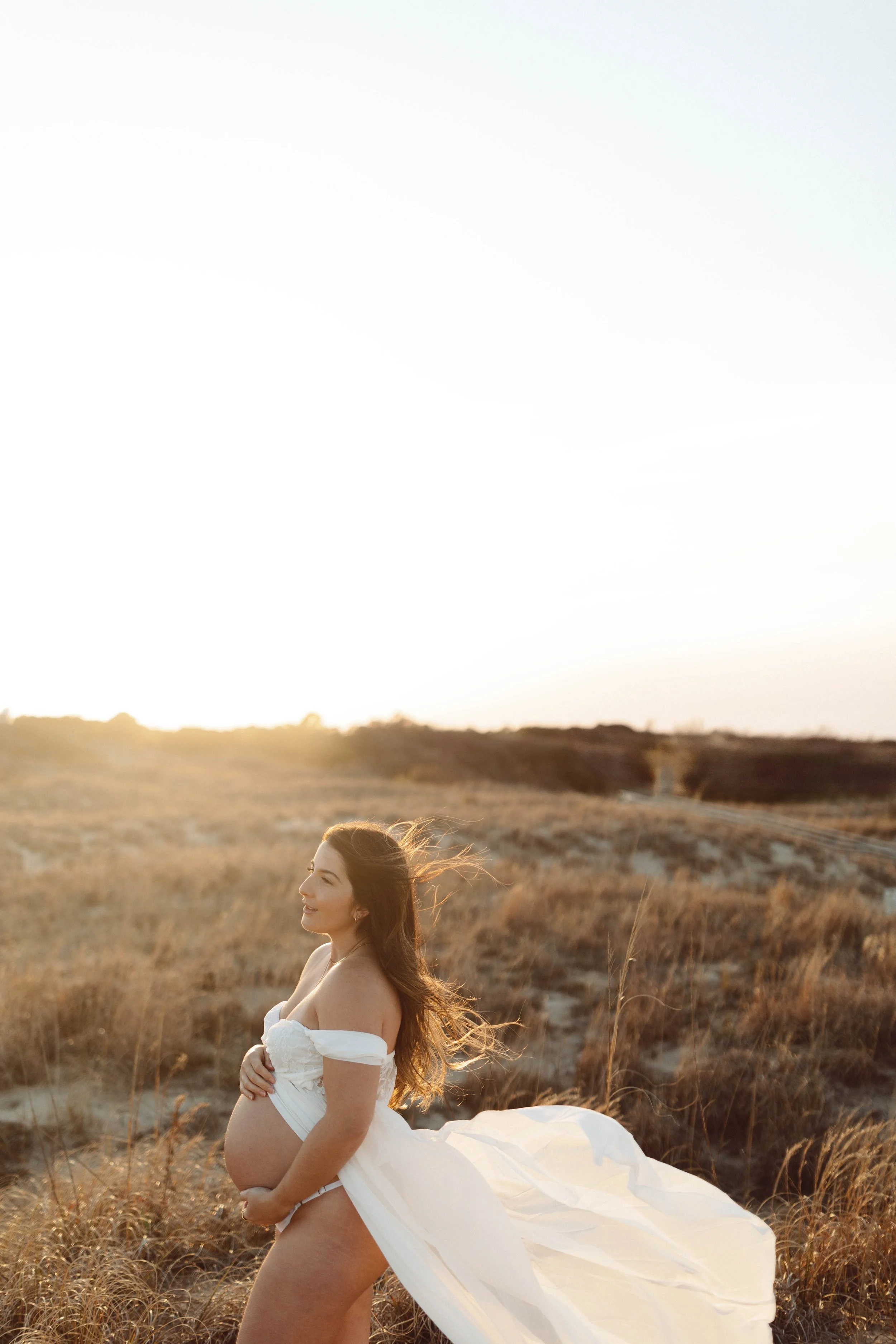 woman stands on golden dunes at Virginia Beach oceanfront cupping pregnant belly while white sheer flowy gown flies around her.