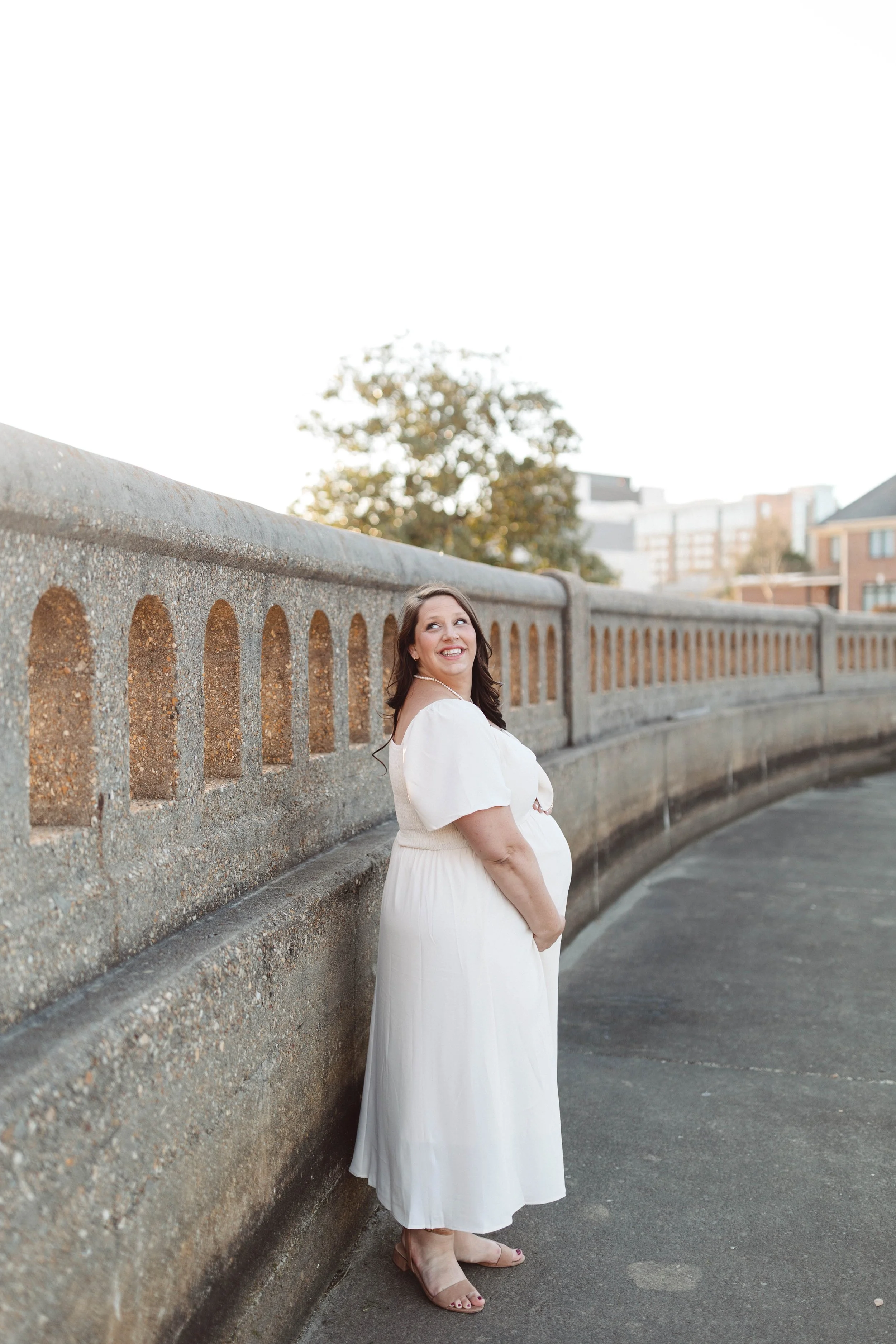 woman stands beside river and granite walkway cupping belly and staring over her shoulder