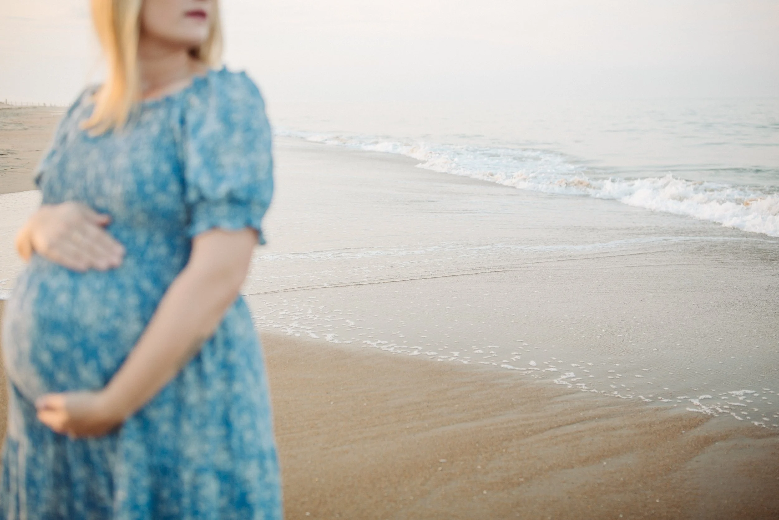 Blurred woman in a blue dress standing on a sandy beach near the ocean, facing away from the camera.