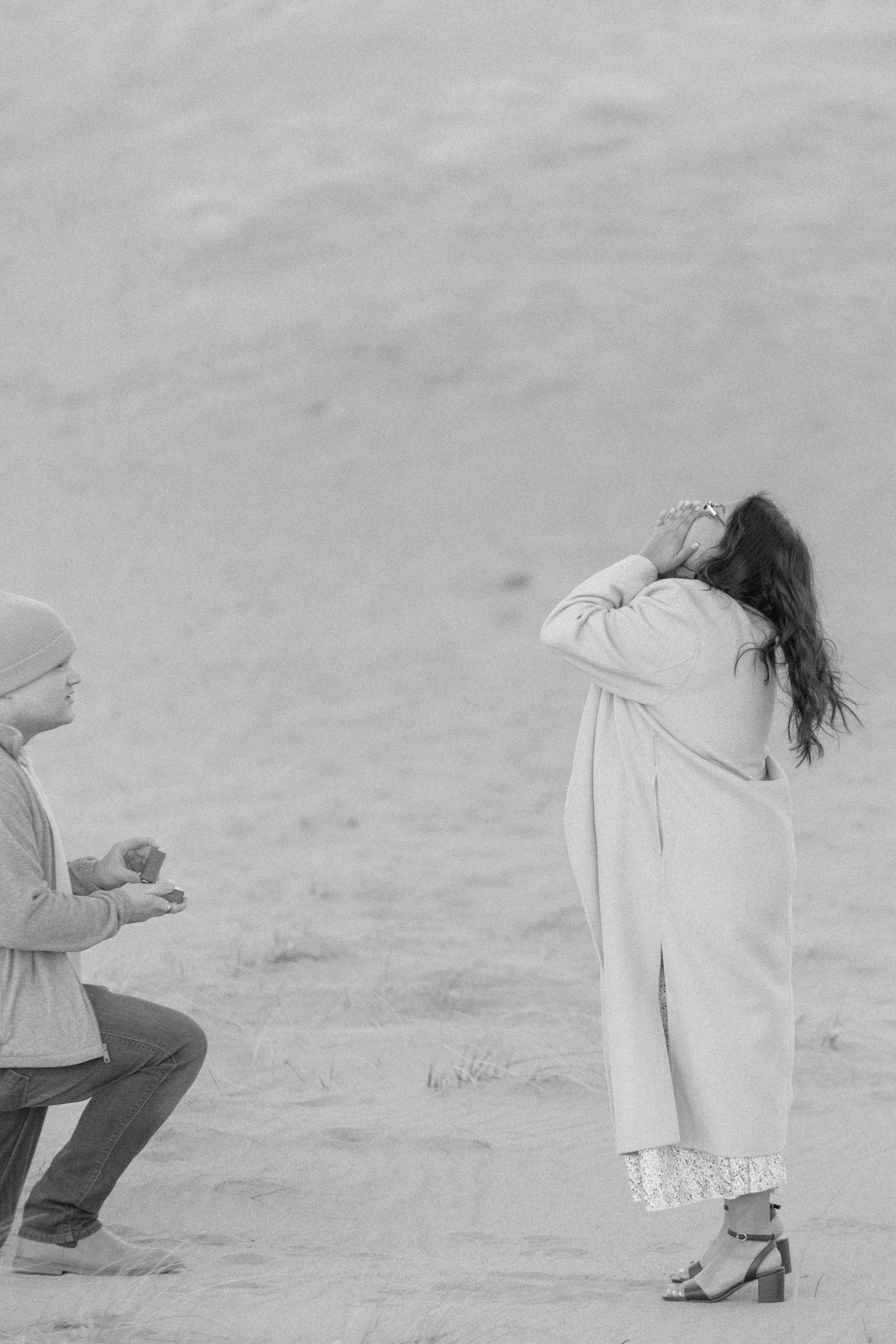 man is down on one knee proposing while woman grabs her mouth in surprise and looks to the sky