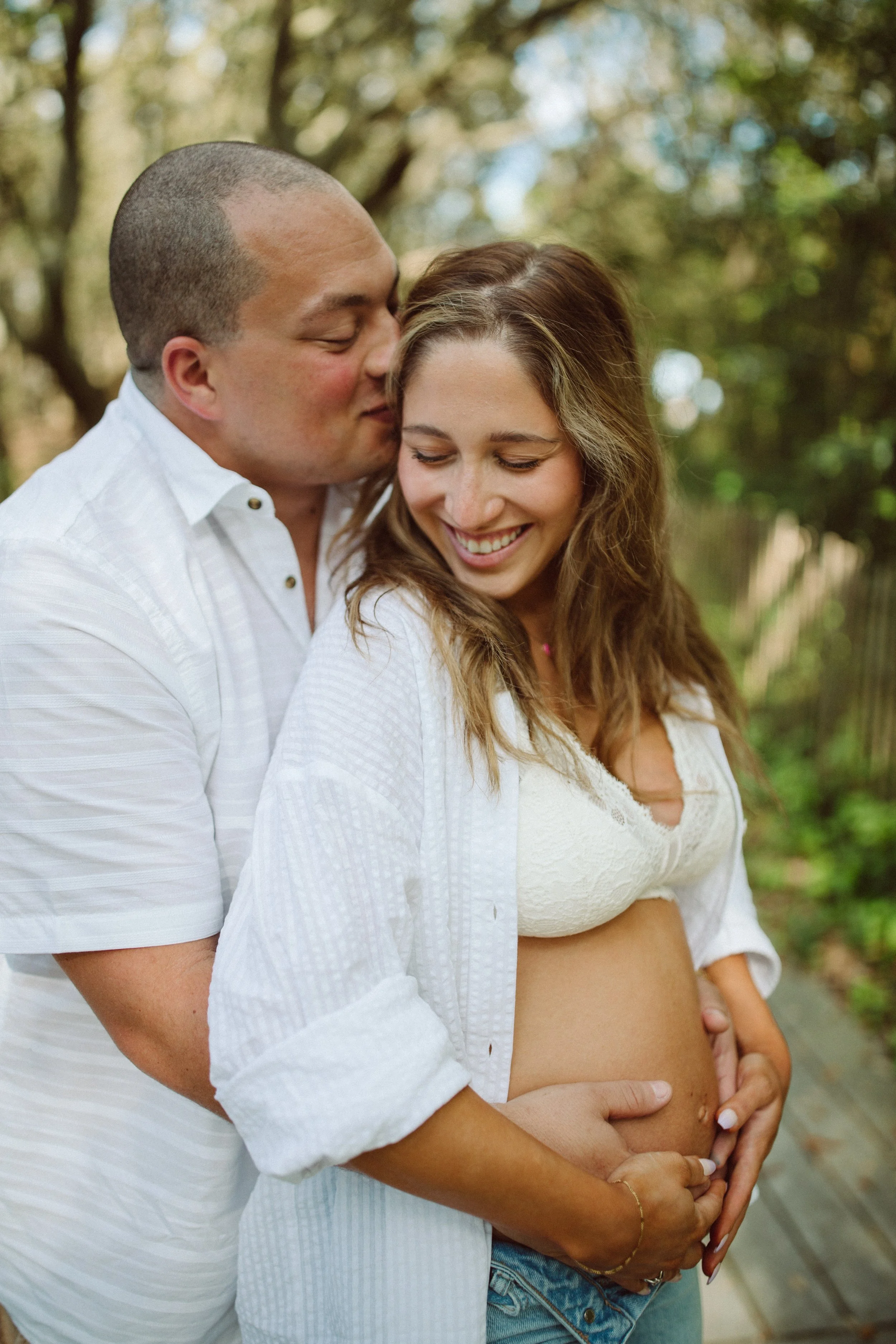 A man and a pregnant woman smiling and embracing outdoors, with the man's face close to the woman's cheek, in a serene, tree-lined setting.