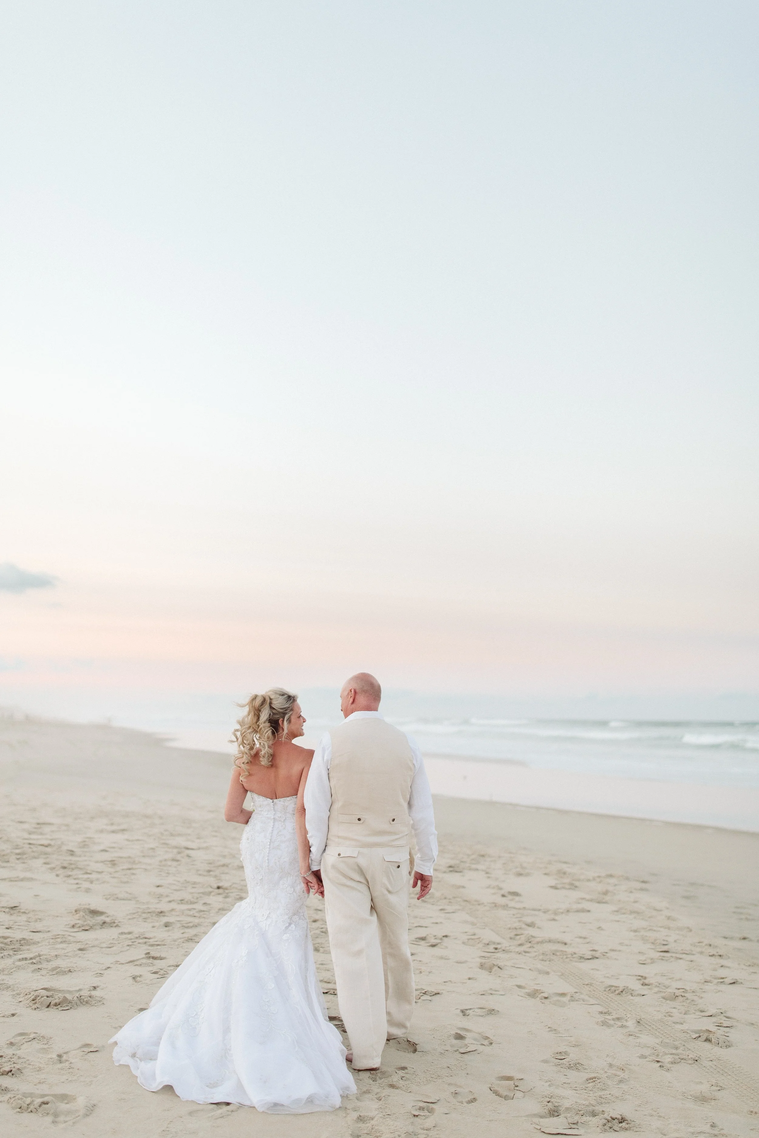 A bride and groom walking hand-in-hand on the beach during sunset.