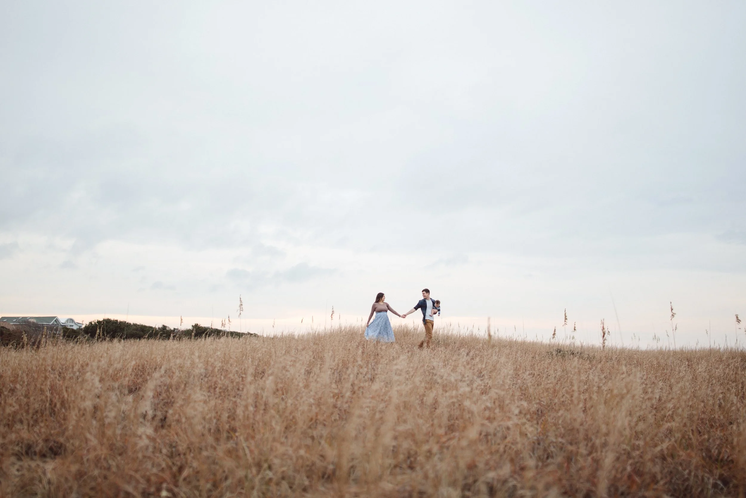 A couple holding hands in a field, with the woman wearing a blue dress and the man wearing a jacket, under a cloudy sky.