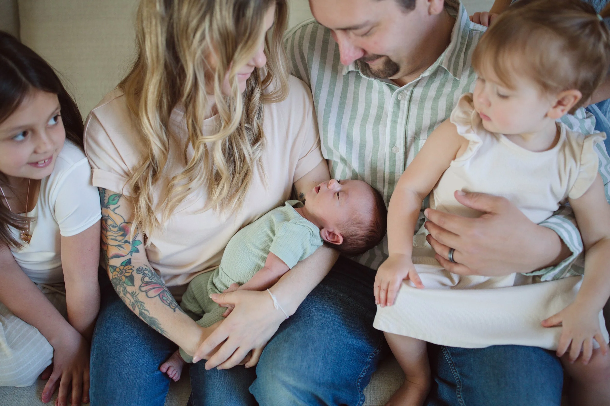 Family gathered together, with a woman holding a newborn baby on her lap, a man sitting beside her, and two young girls sitting nearby, all looking at the baby.