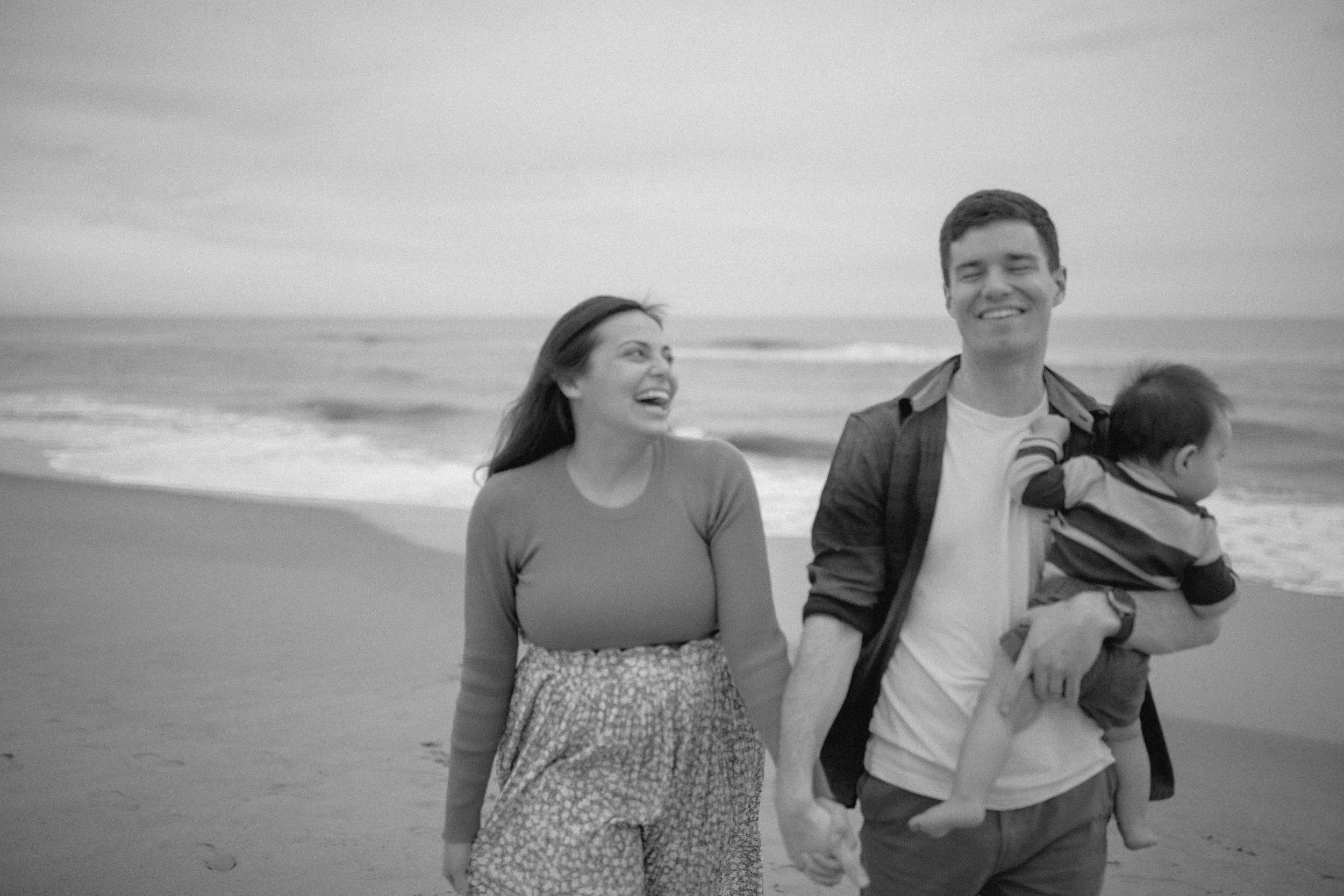 A smiling couple holding hands and walking along a beach with a toddler in a carrier on the man's chest, ocean waves in the background, in black and white.