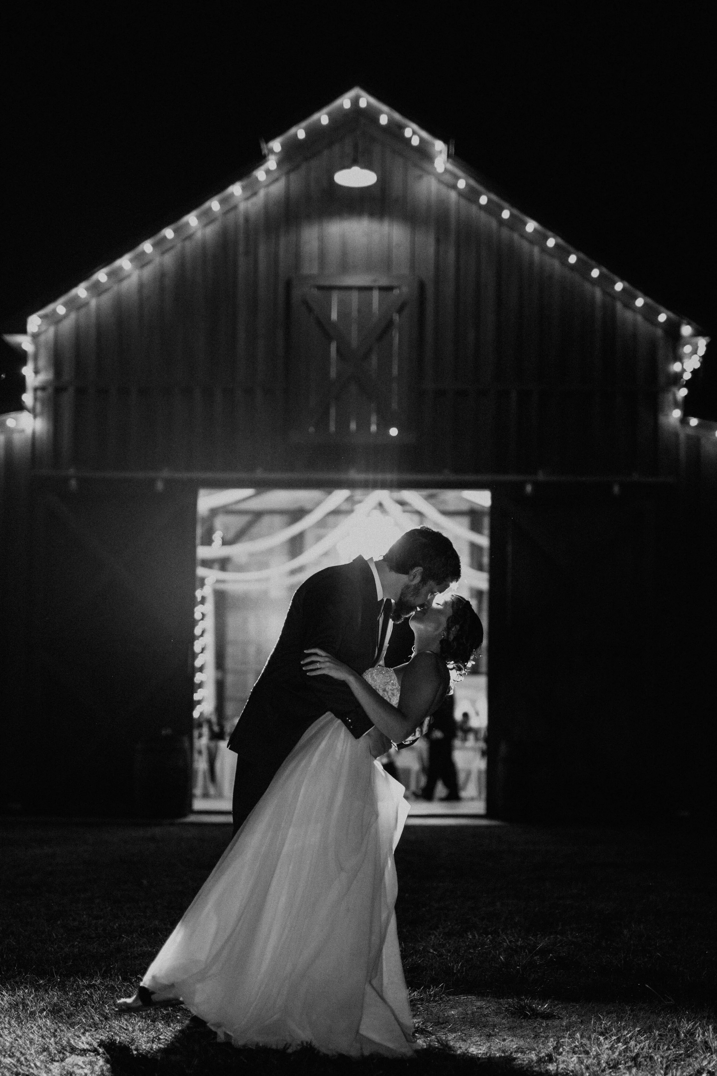 A black and white photo of a newlywed couple dancing outside a barn at night, with the barn decorated with string lights.