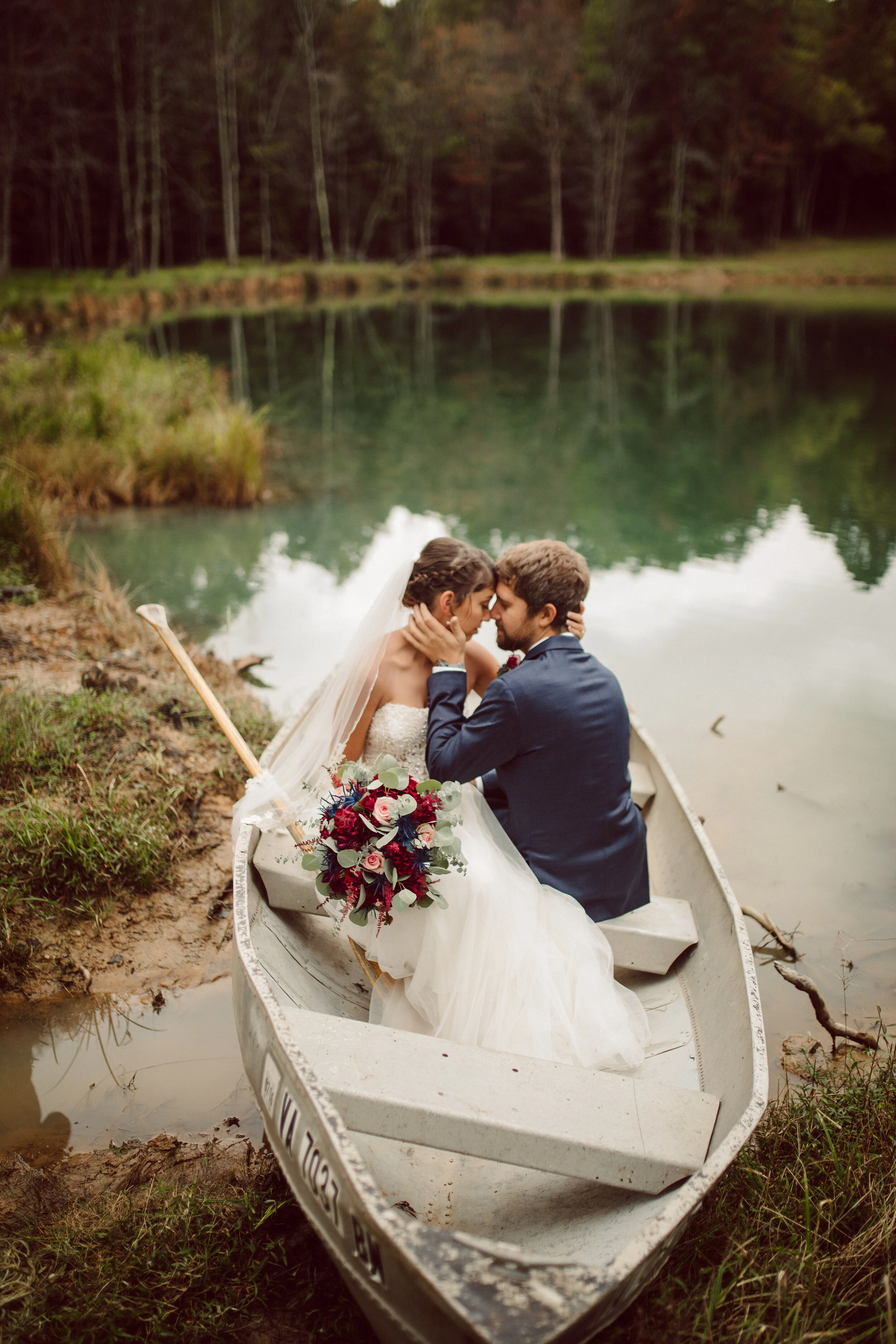A bride and groom sitting in a small rowboat on a lake, touching foreheads, with the bride holding a bouquet of red, white, and pink flowers, surrounded by nature.