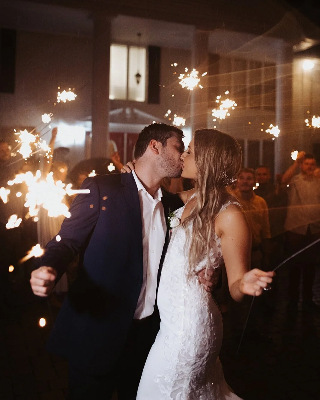 A bride and groom kissing at their wedding reception surrounded by sparklers and guests.