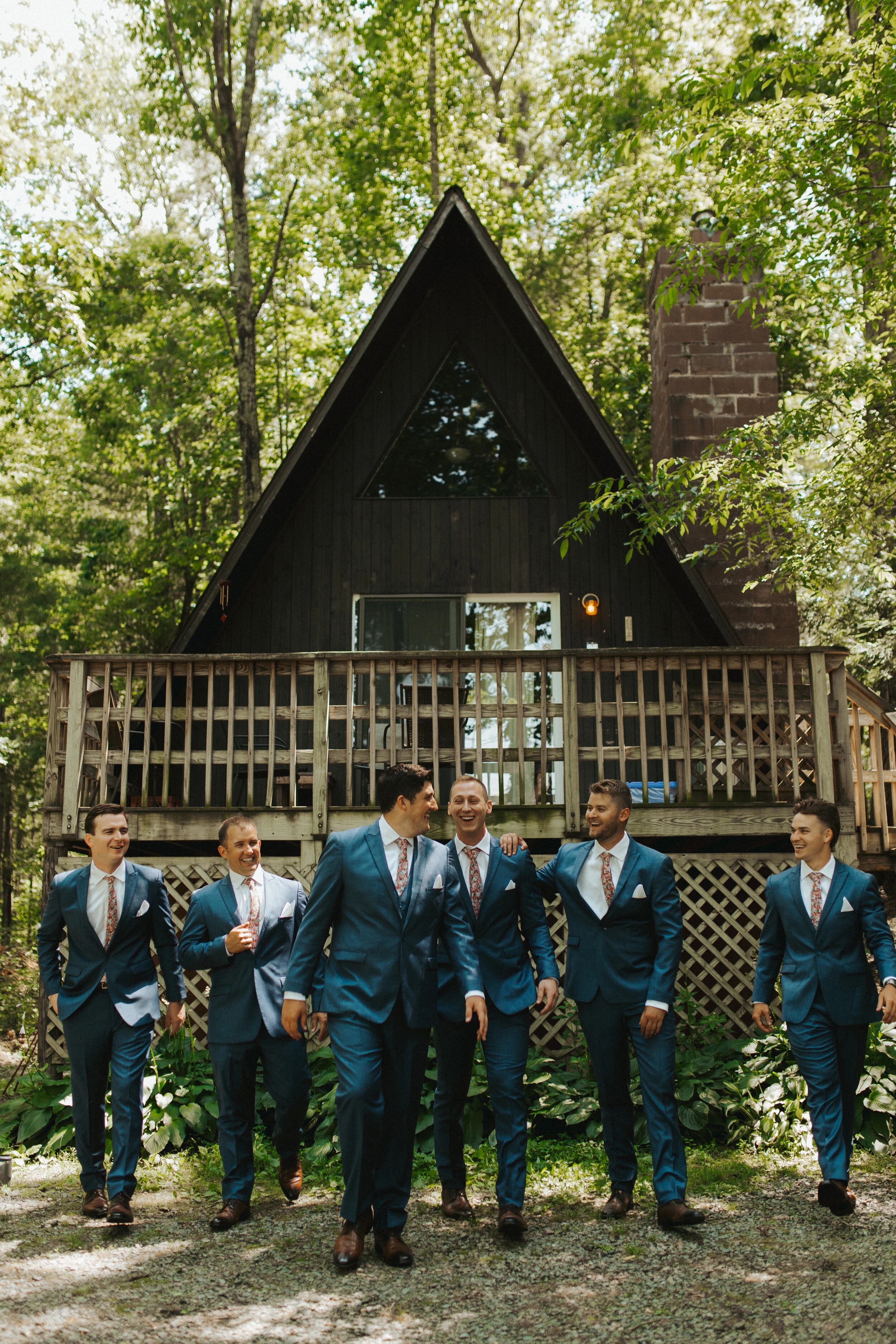 A group of six men in matching blue suits and floral ties walking and laughing outdoors in front of a black A-frame house with a wooden balcony and green trees.