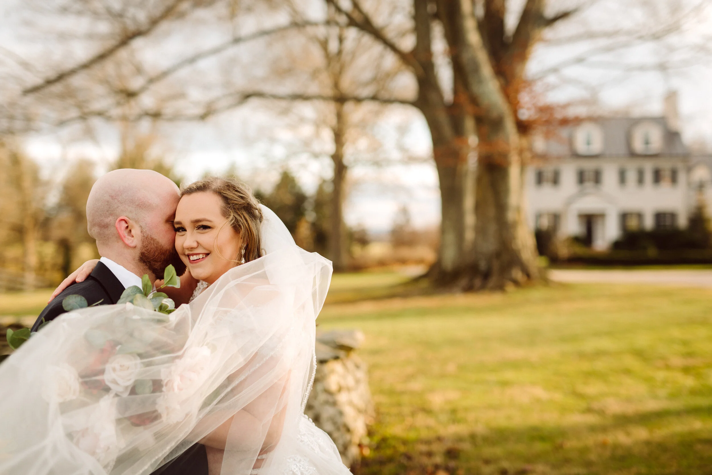 A bride and groom embracing outdoors near a large tree and a white house. The bride is smiling and holding a bouquet, while the groom whispers in her ear. Fall foliage is visible in the background.