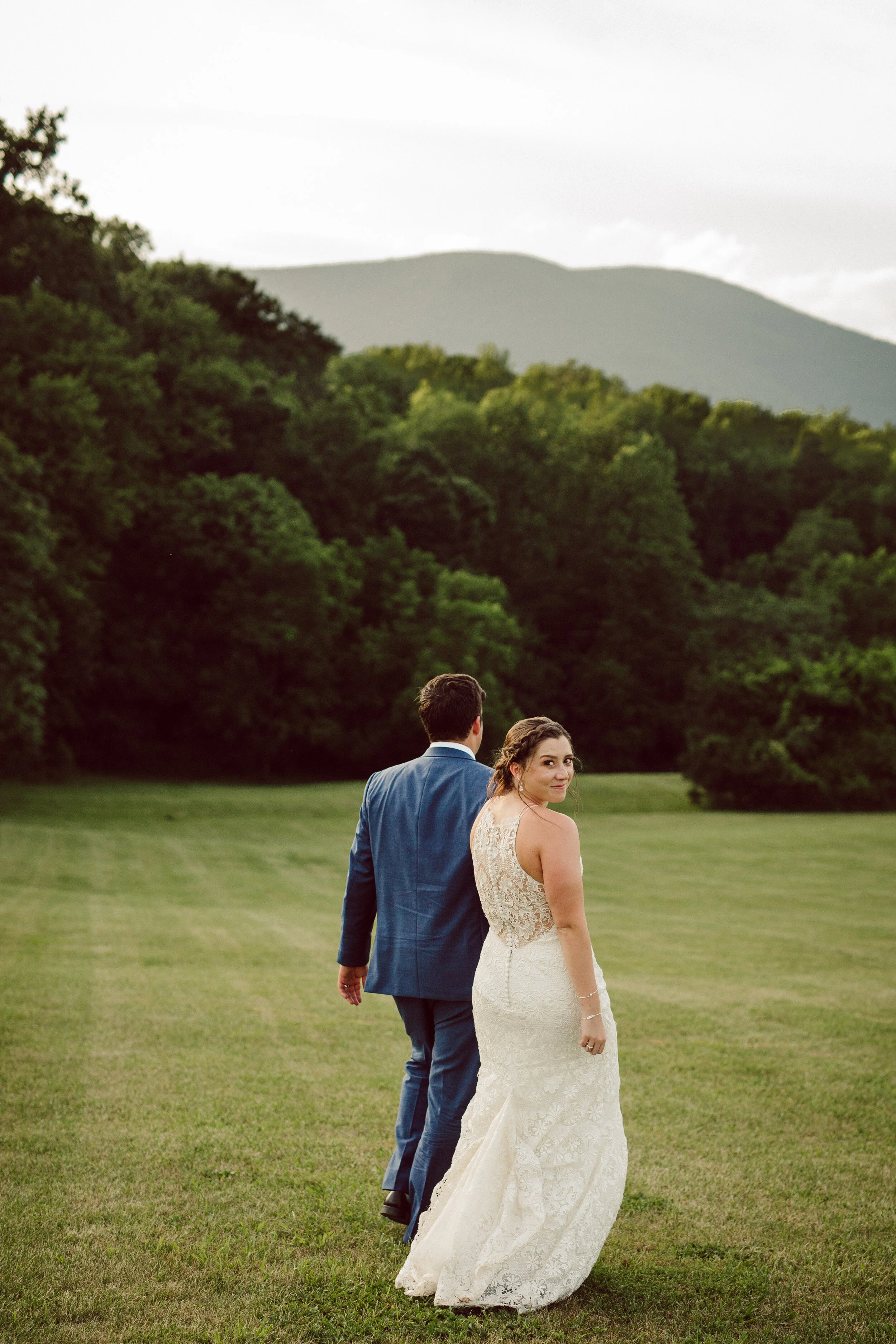 Bride in a white wedding dress looking over her shoulder, groom in a blue suit walking away on a grassy field with trees and mountains in the background.