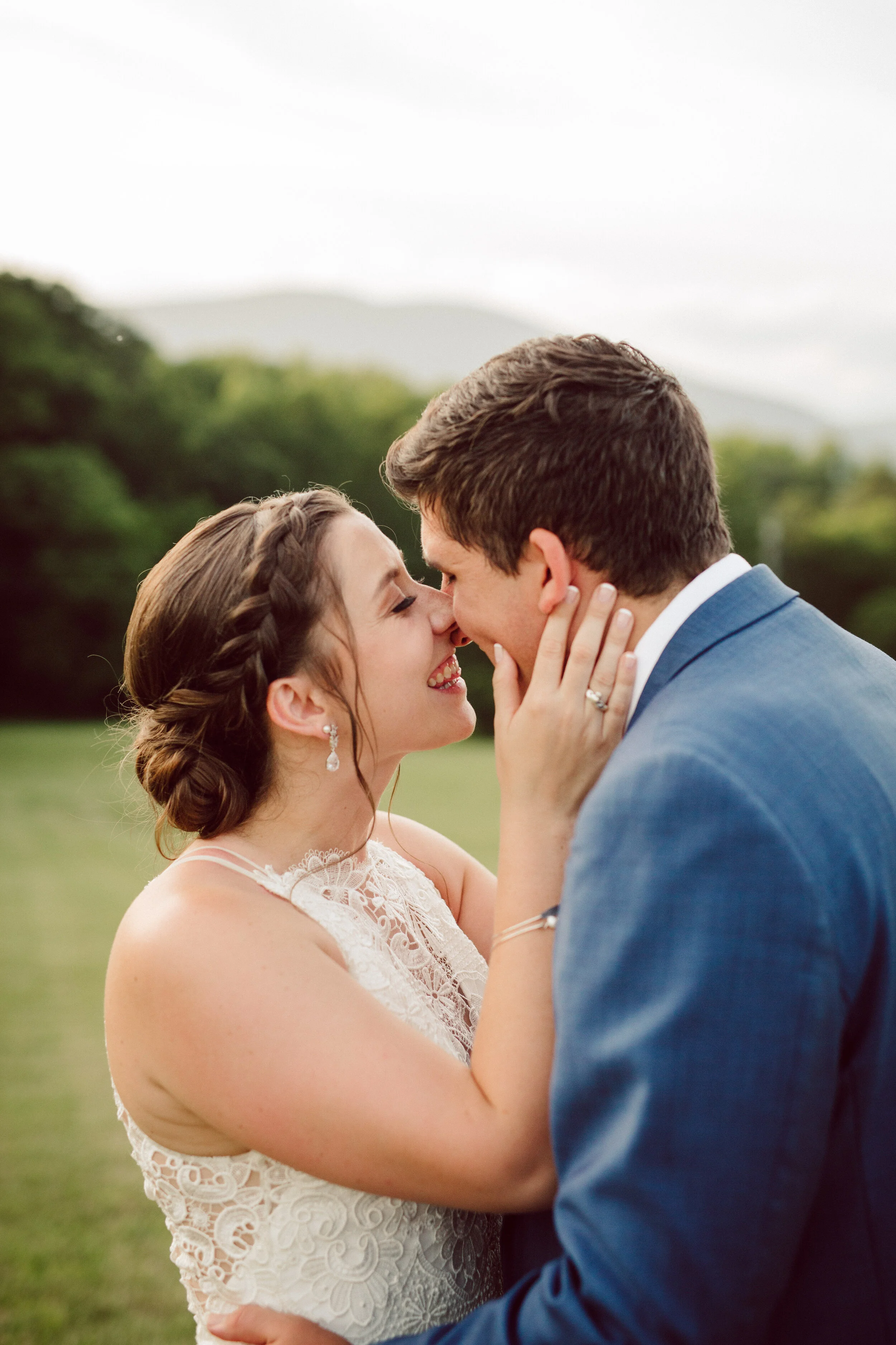 A bride and groom in an outdoor setting, smiling and touching noses, with the bride holding the groom's face and the groom's hand around her waist, during a wedding celebration.