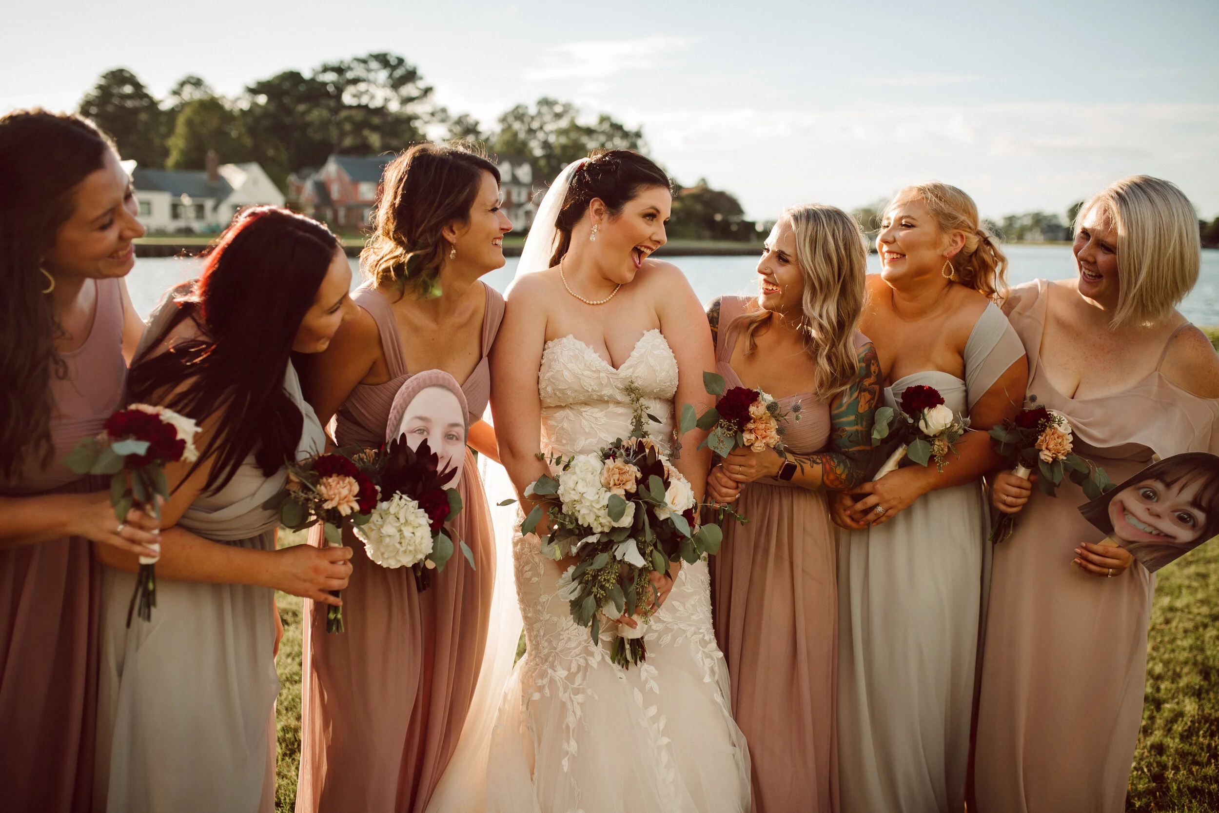 Bride in a white wedding dress surrounded by six bridesmaids holding bouquets, outdoors with water and houses in the background, all smiling and laughing.