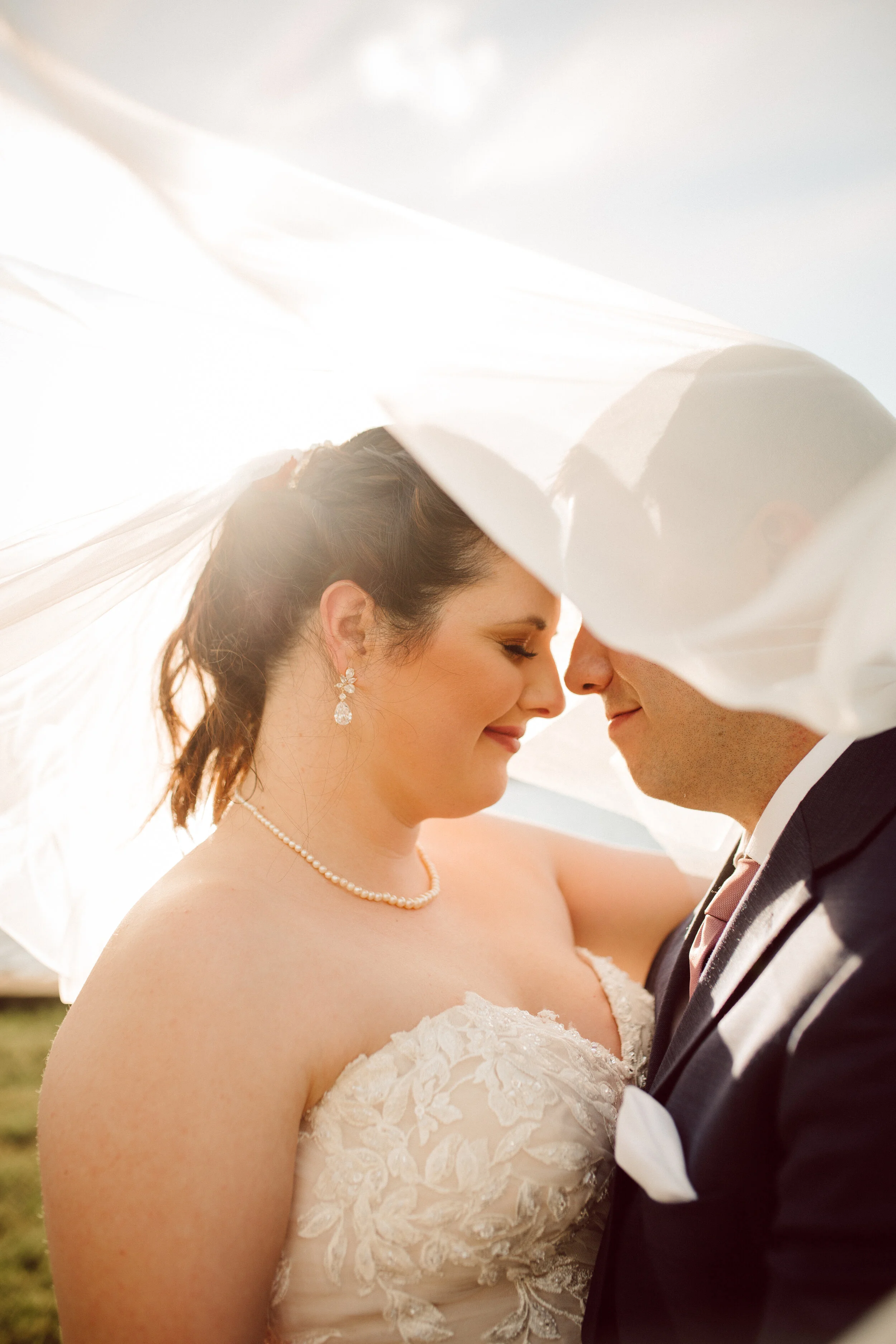 A bride and groom on their wedding day, smiling and touching foreheads under a white veil, outdoors with sunlight.