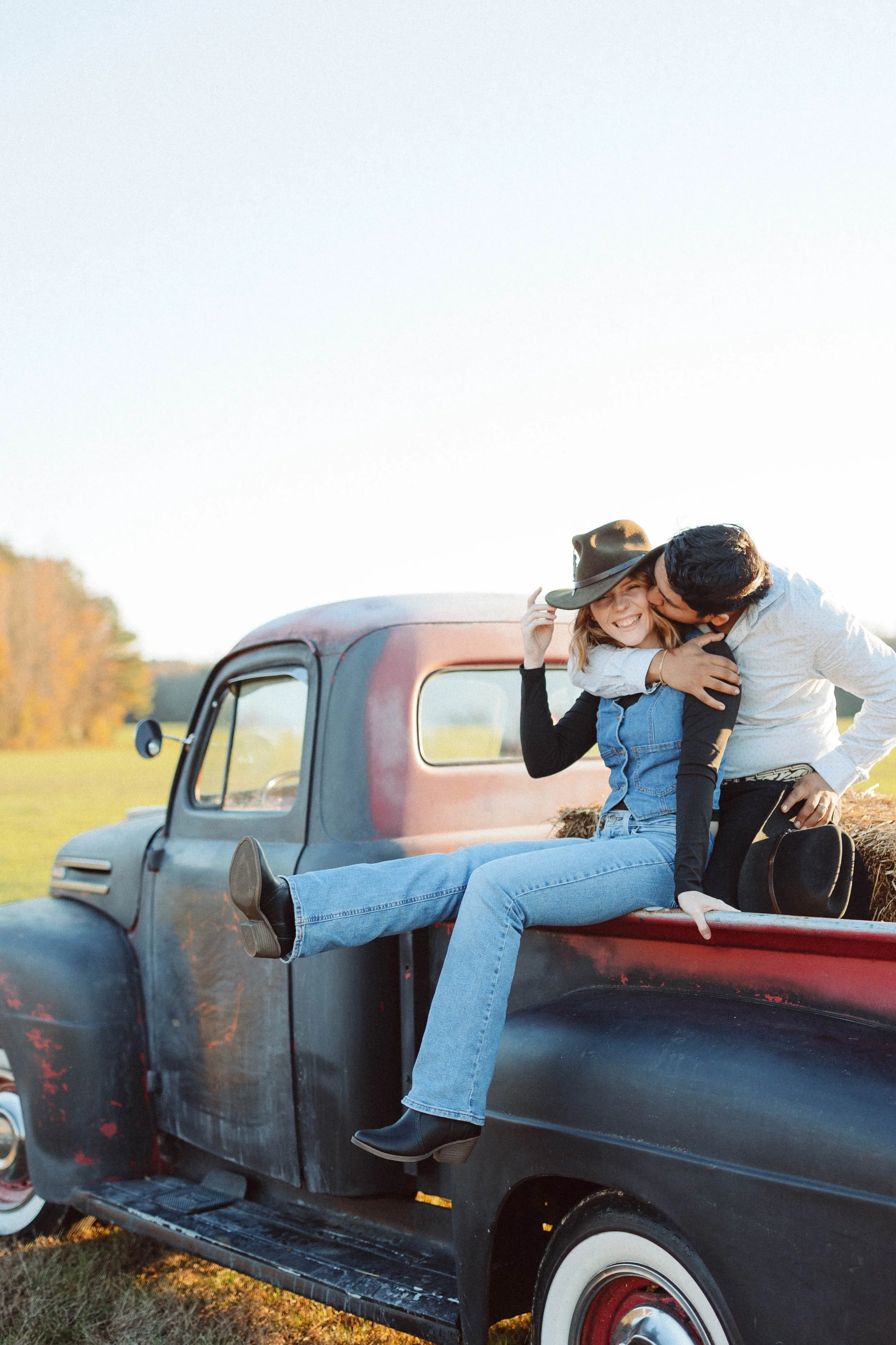 Couple sits on top of vintage ford pickup truck wearing western attire and looking into the distance
