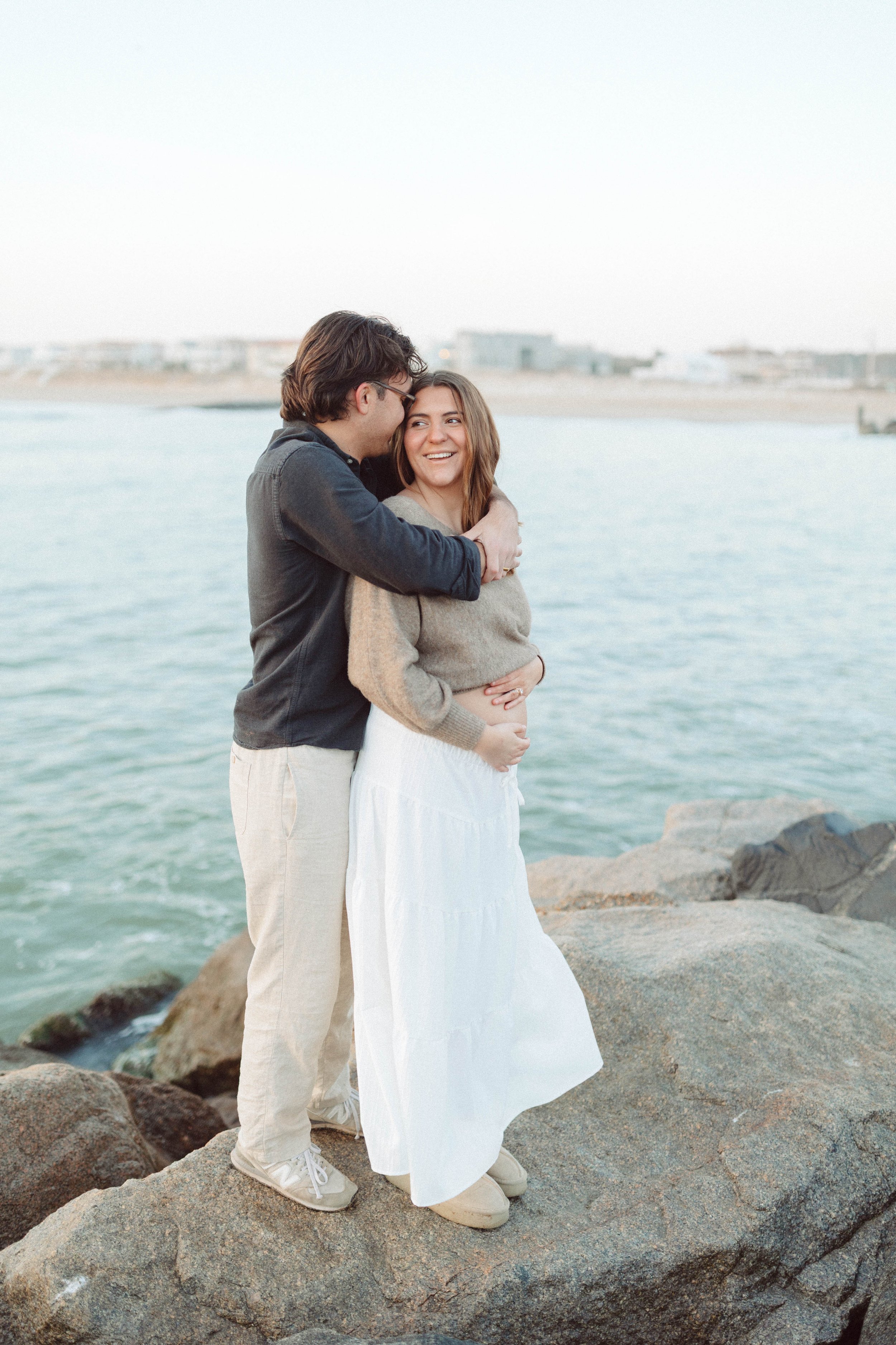 couple stands on a rocky embankment embracing each other and laughing while holding a strip of ultrasound pictures.