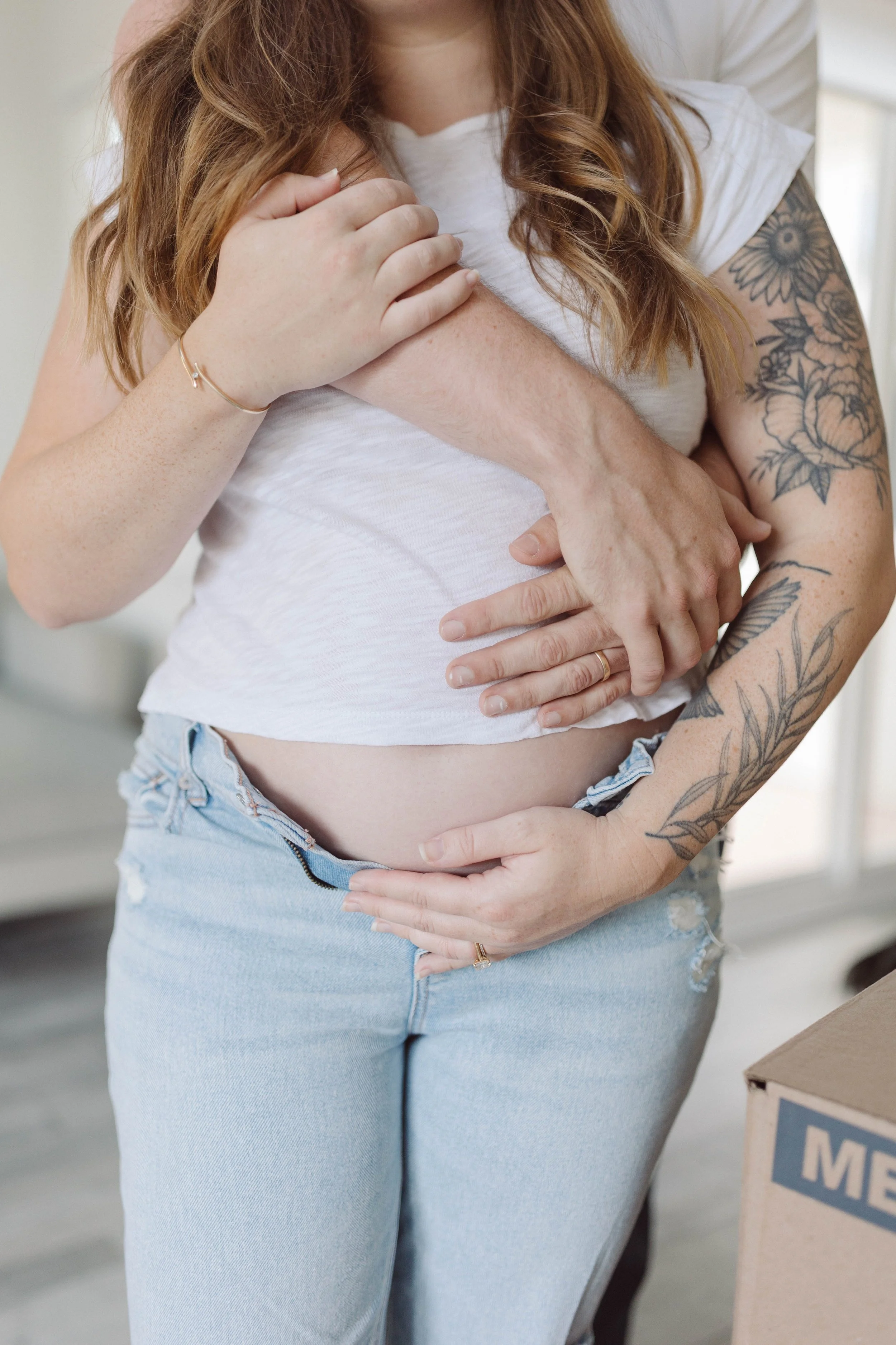 Close-up of a pregnant woman with red hair and tattoos on her arm, holding her belly with her hand, while another person's hand is on her shoulder.