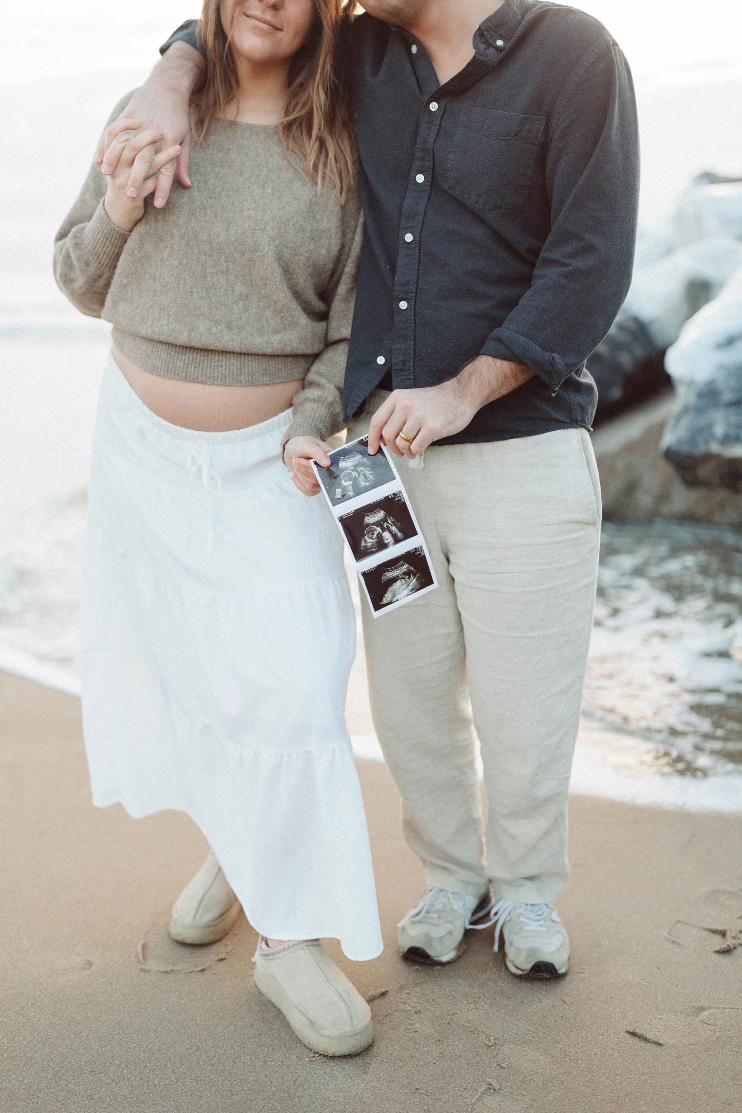 woman and man standing side by side with a rocky beach in the background holding ultrasound pictures