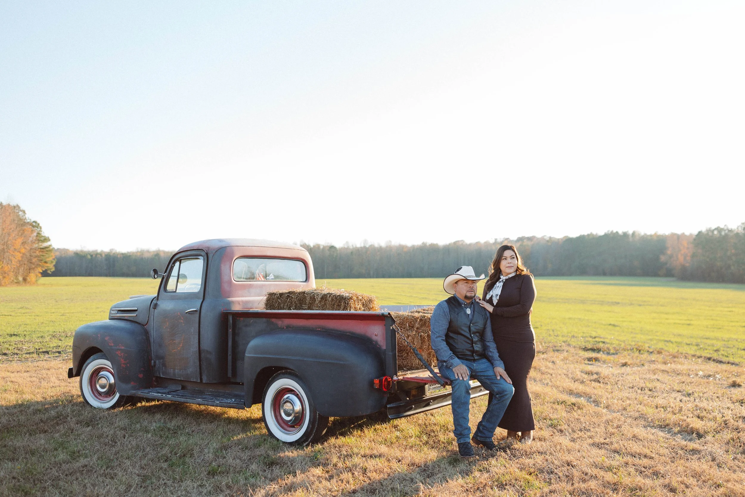 couple stands next to vintage antique ford pickup truck gazing at the horizon while wearing western attire and standing in a field