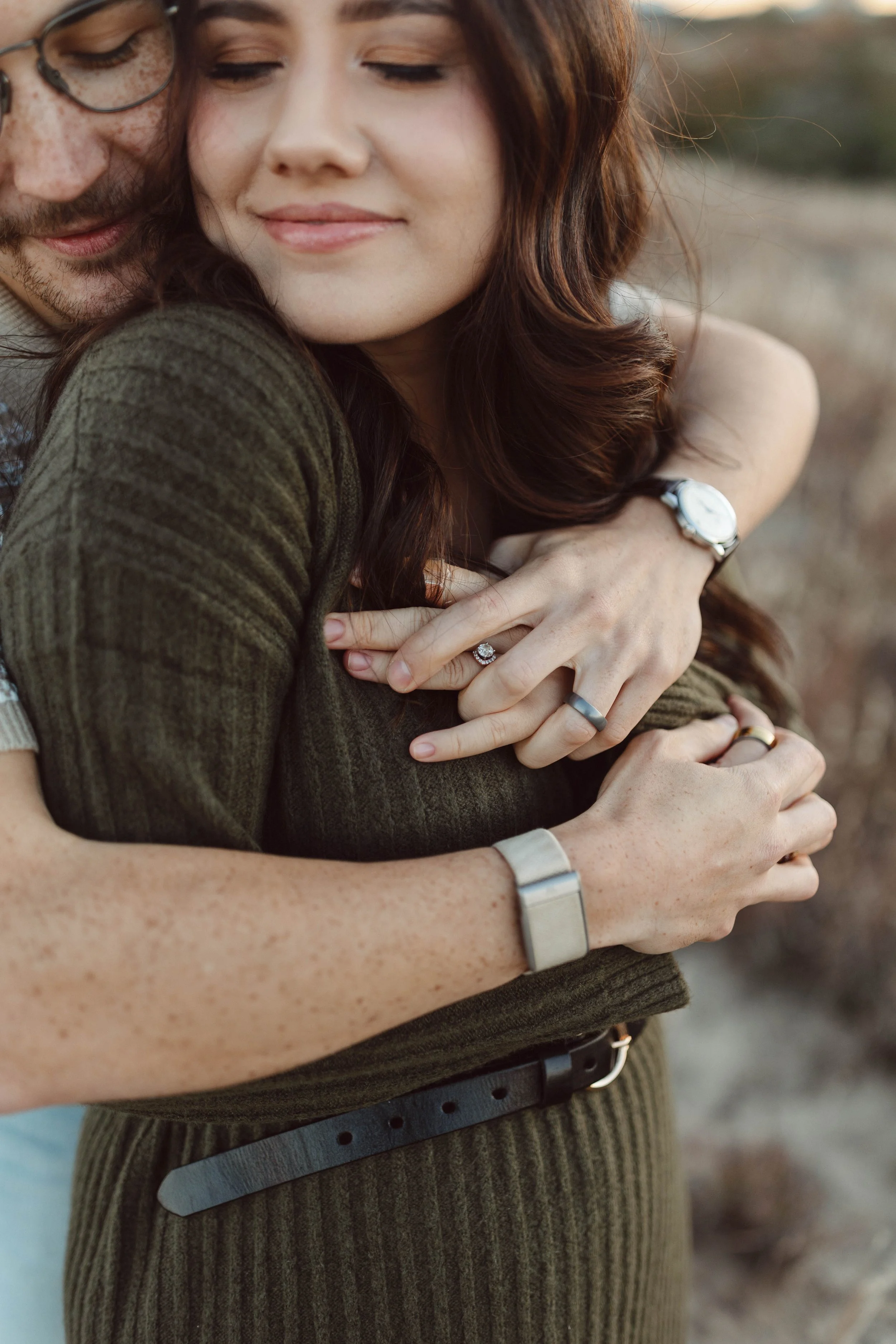 A couple embracing outdoors with women displaying an engagement ring, both wearing wristwatches and rings, during sunset.