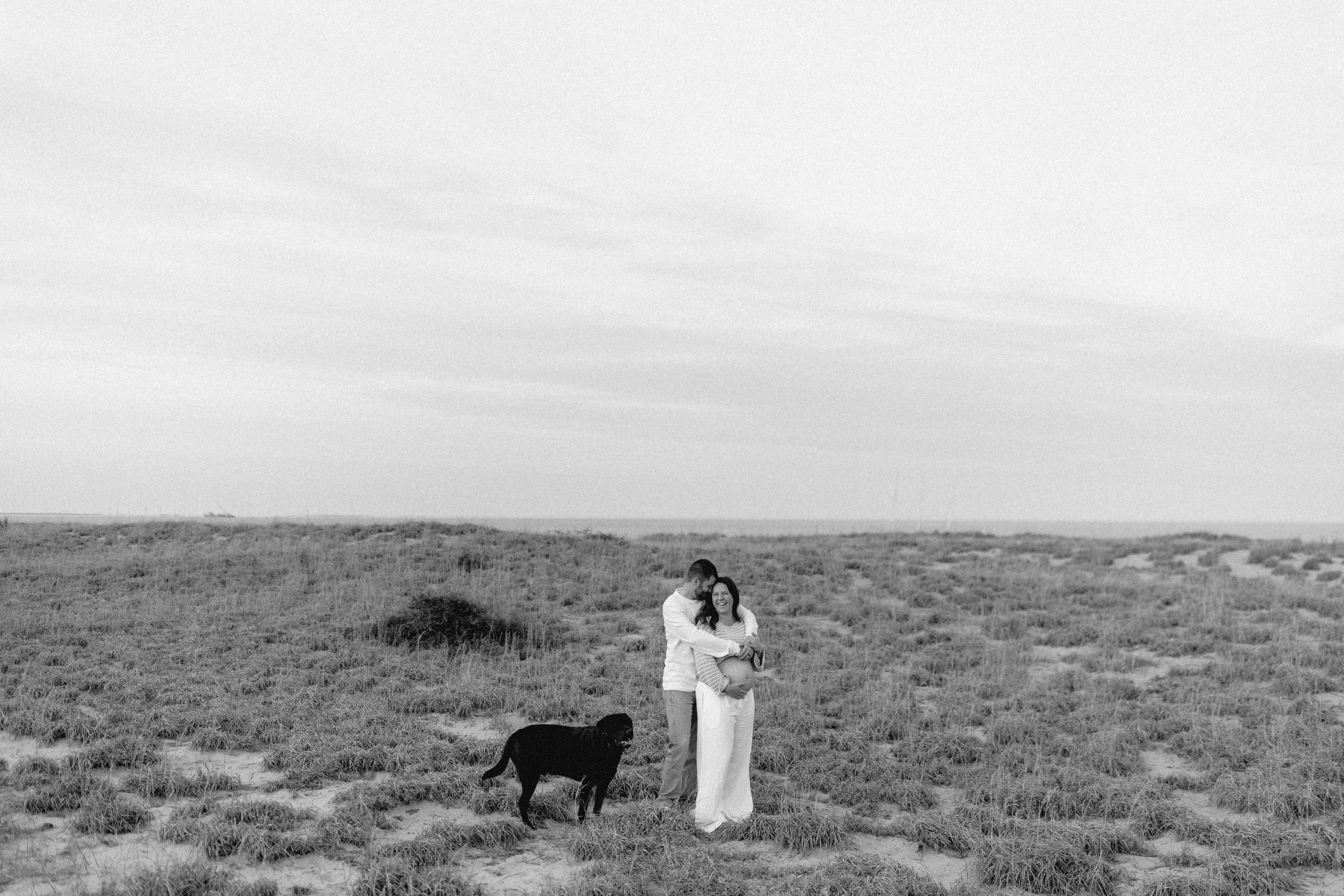 couple stands on dunes embracing woman's pregnant belly next to black lab dog.