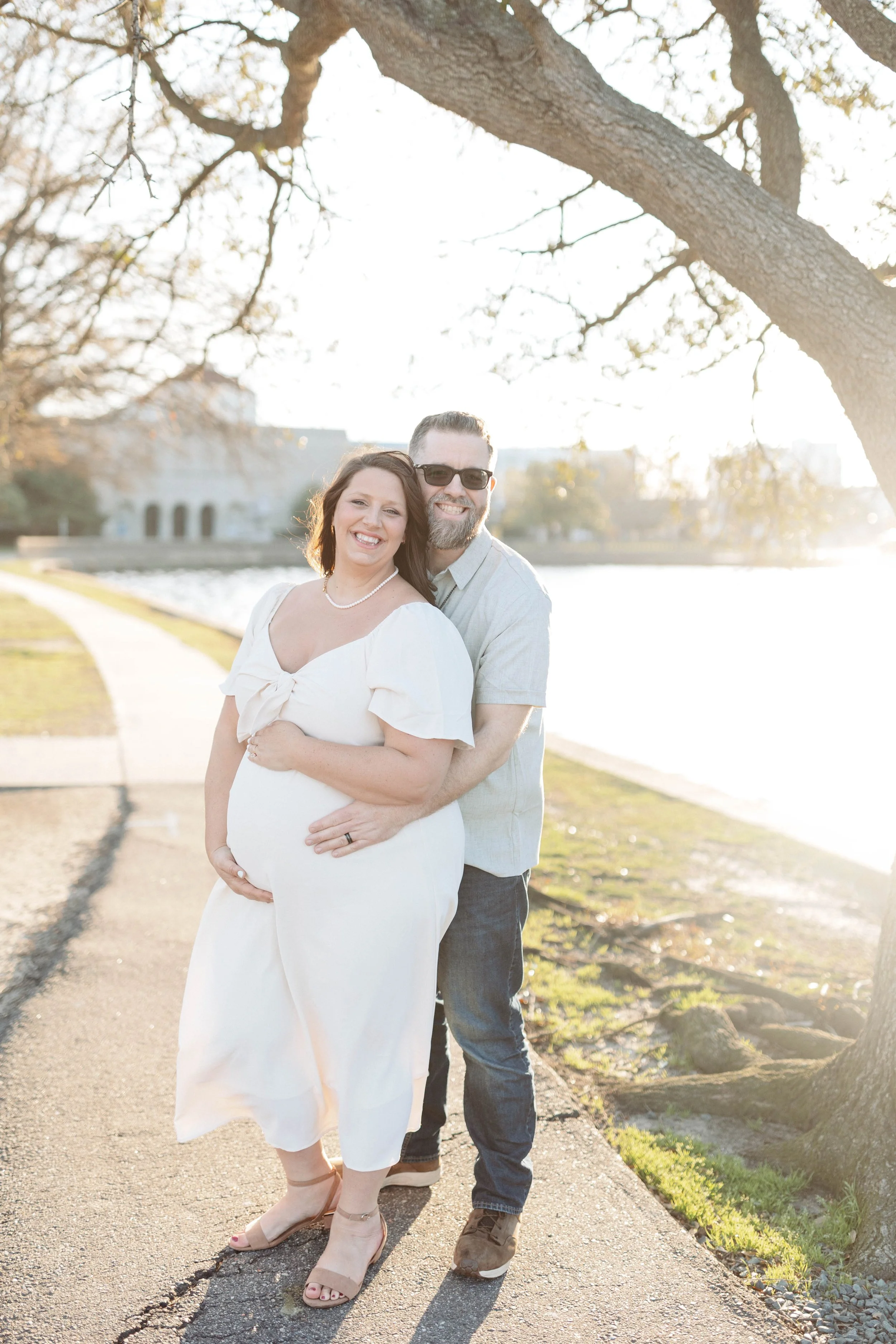 man hugs woman from behind standing alongside river in front of historic museum. woman cups pregnant belly while wearing a white dress and smiling at the camera.