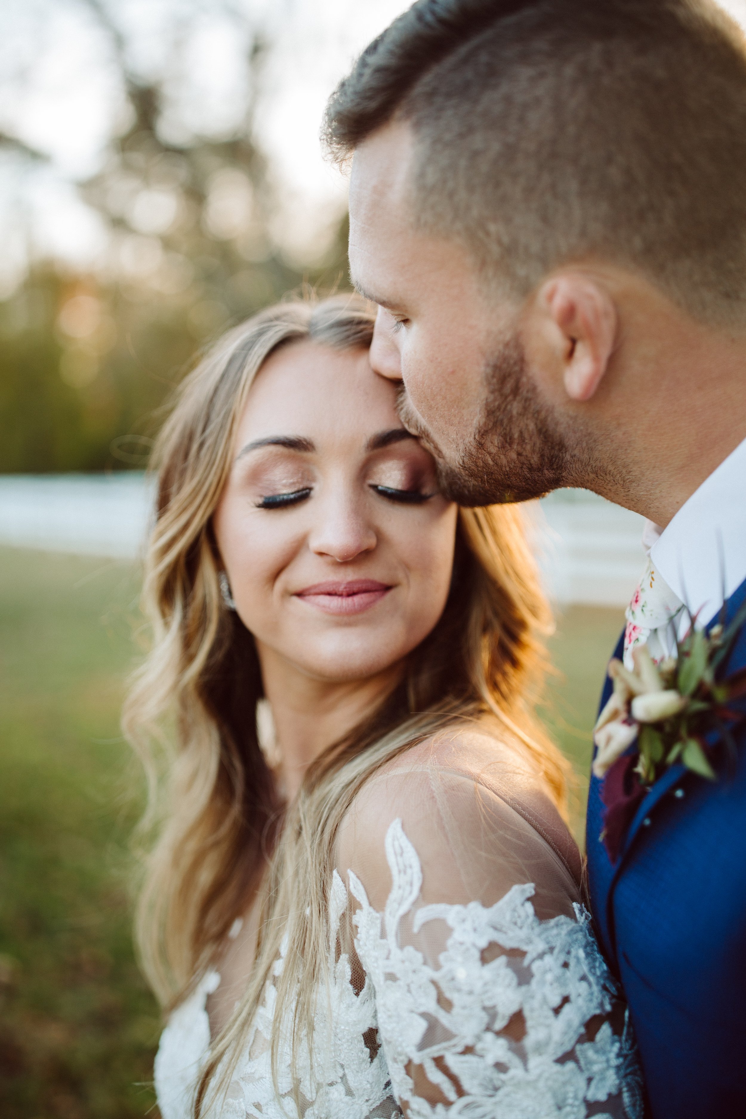 A bride and groom in a close embrace, with the groom kissing the bride's forehead outdoors during sunset.