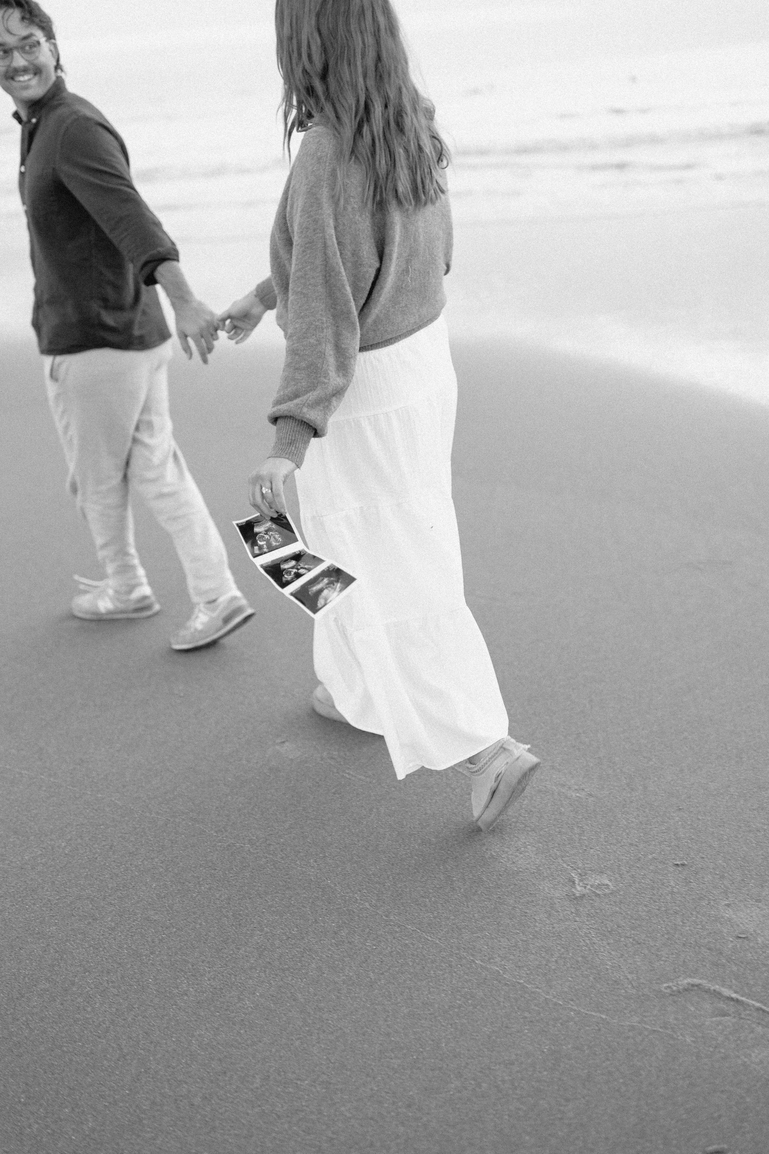black and white picture of couple walking in motion on the beach holding a strip of ultrasound pictures. 