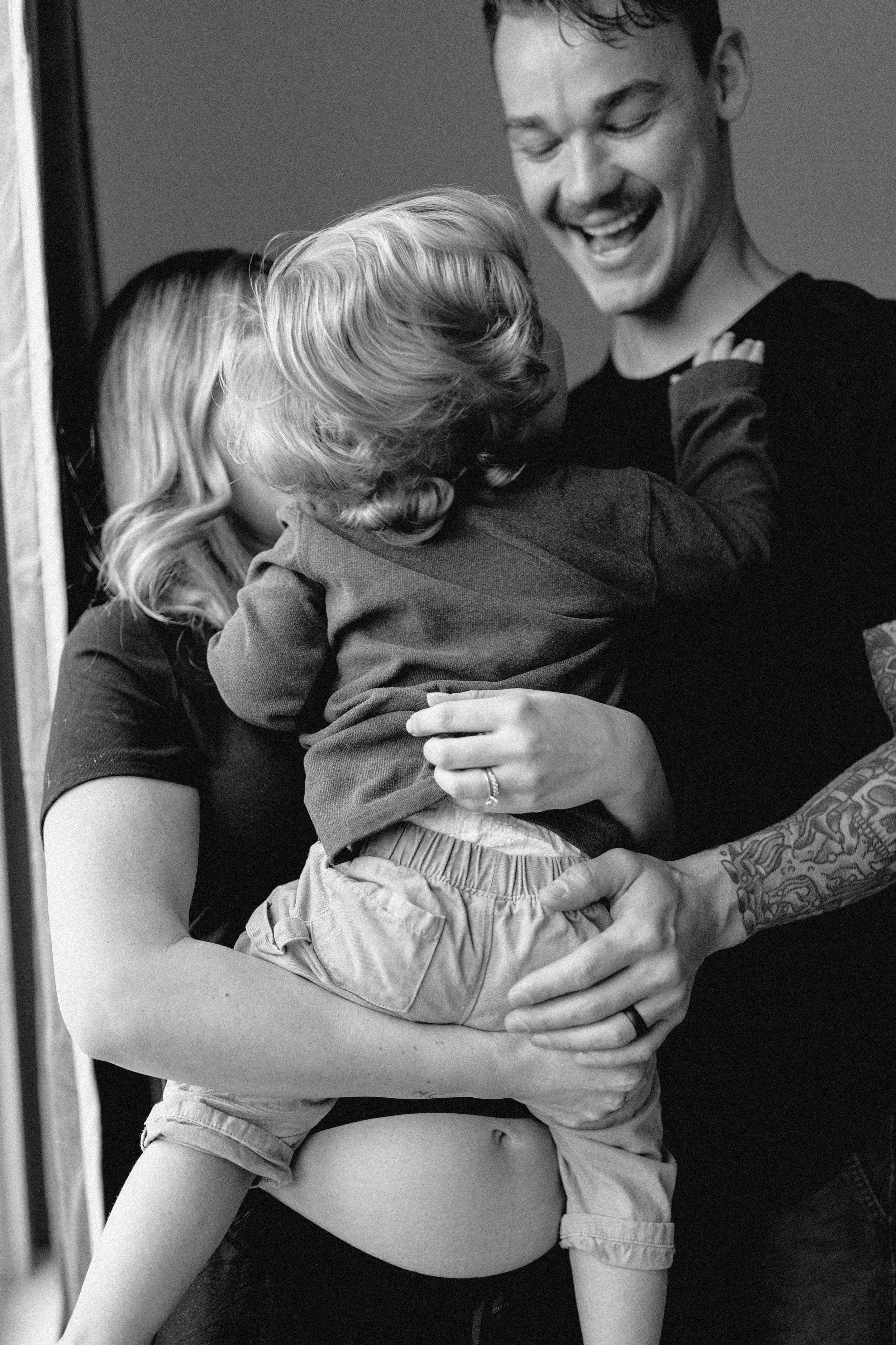 black and white image of tattooed man holding son over wife's pregnant belly while they all smile and laugh